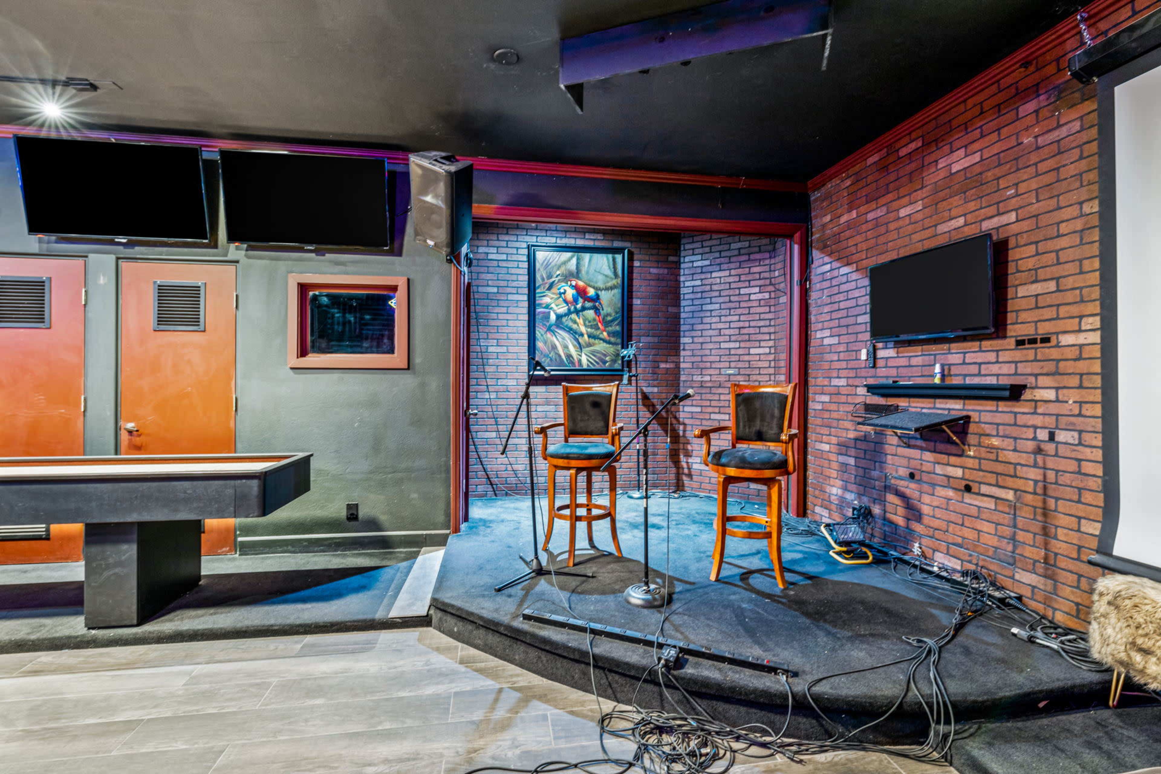 The image shows a small performance stage in a brick-walled venue, featuring two empty chairs, microphones, and several screens mounted on the walls.