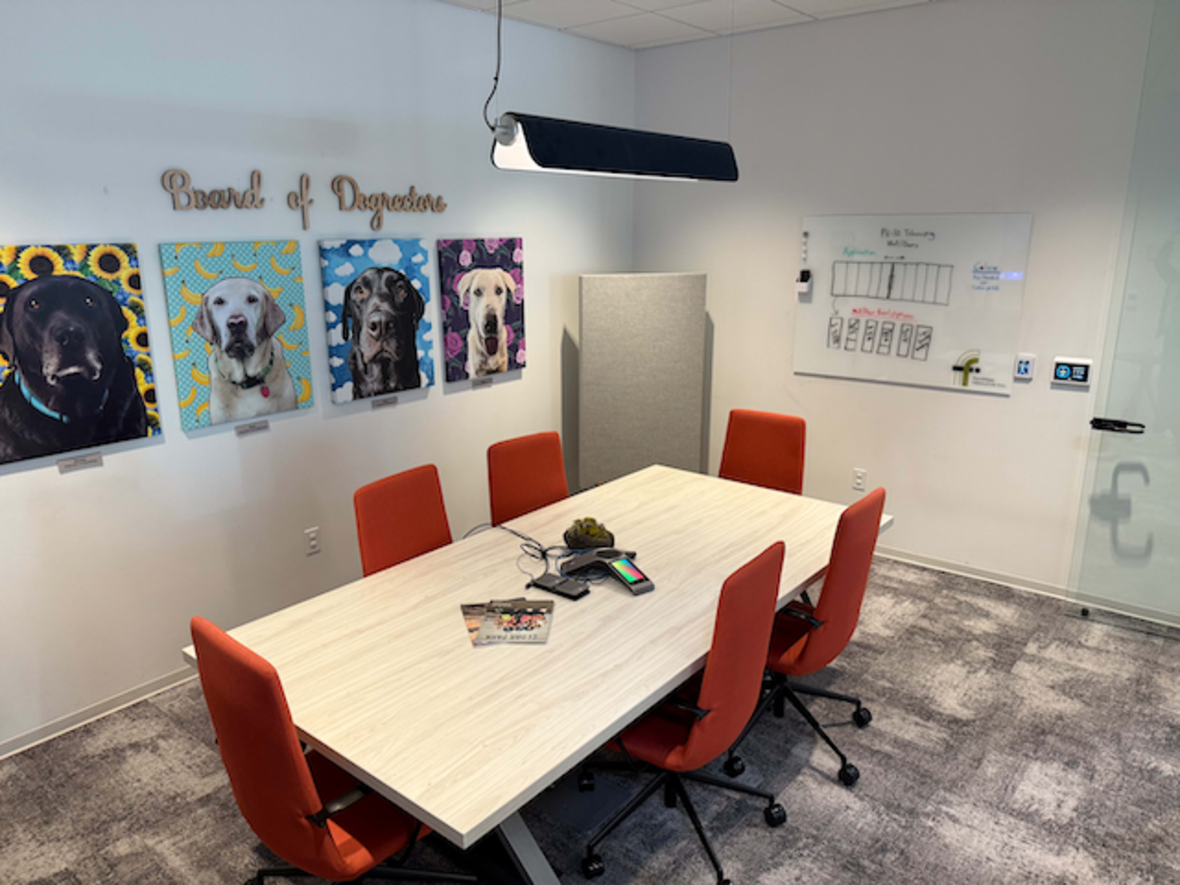 A modern conference room features a large table surrounded by orange chairs, with framed dog portraits on the wall and a whiteboard on another.