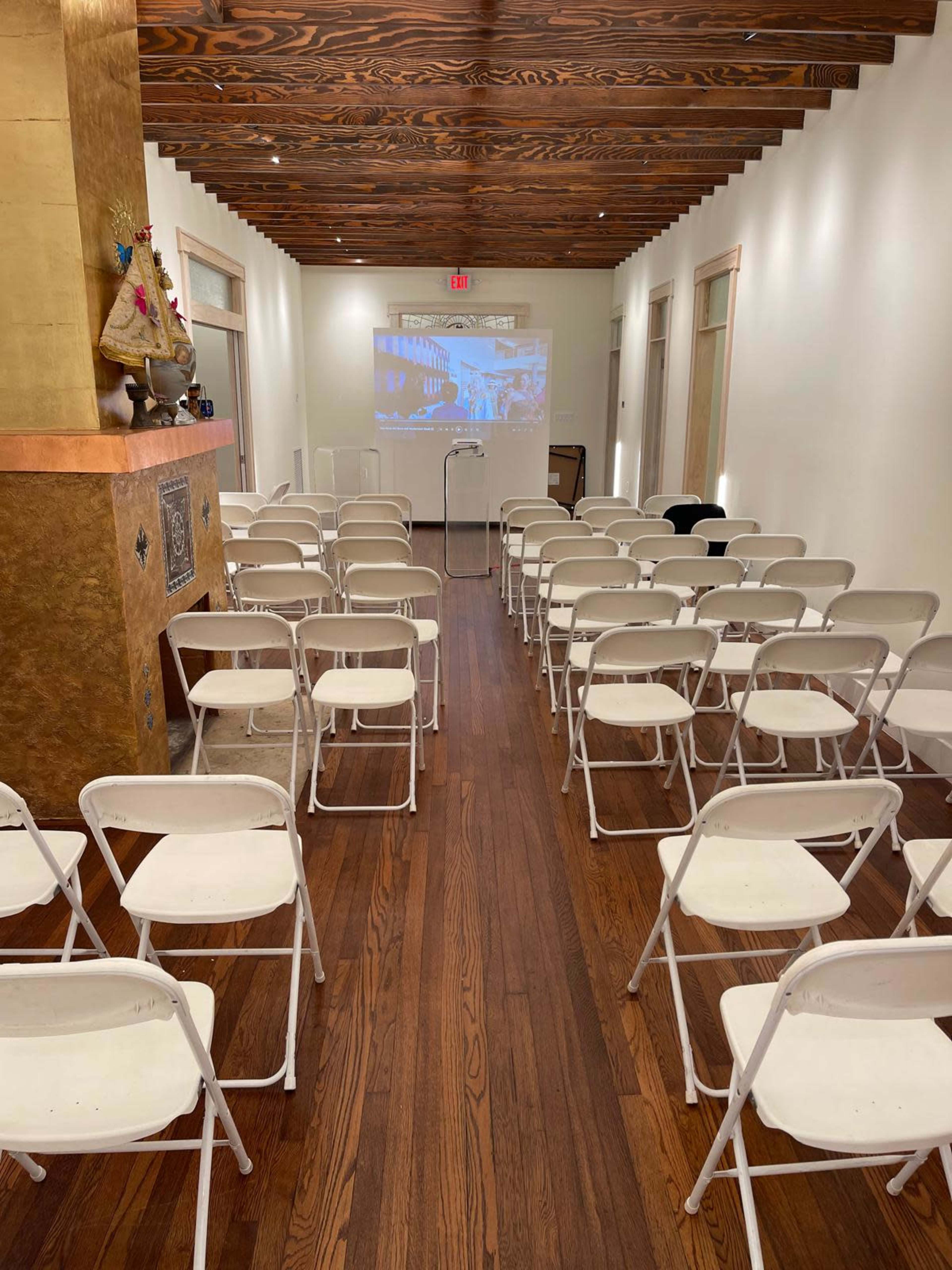 A room with wooden floors and a row of white folding chairs facing a projection screen at one end.