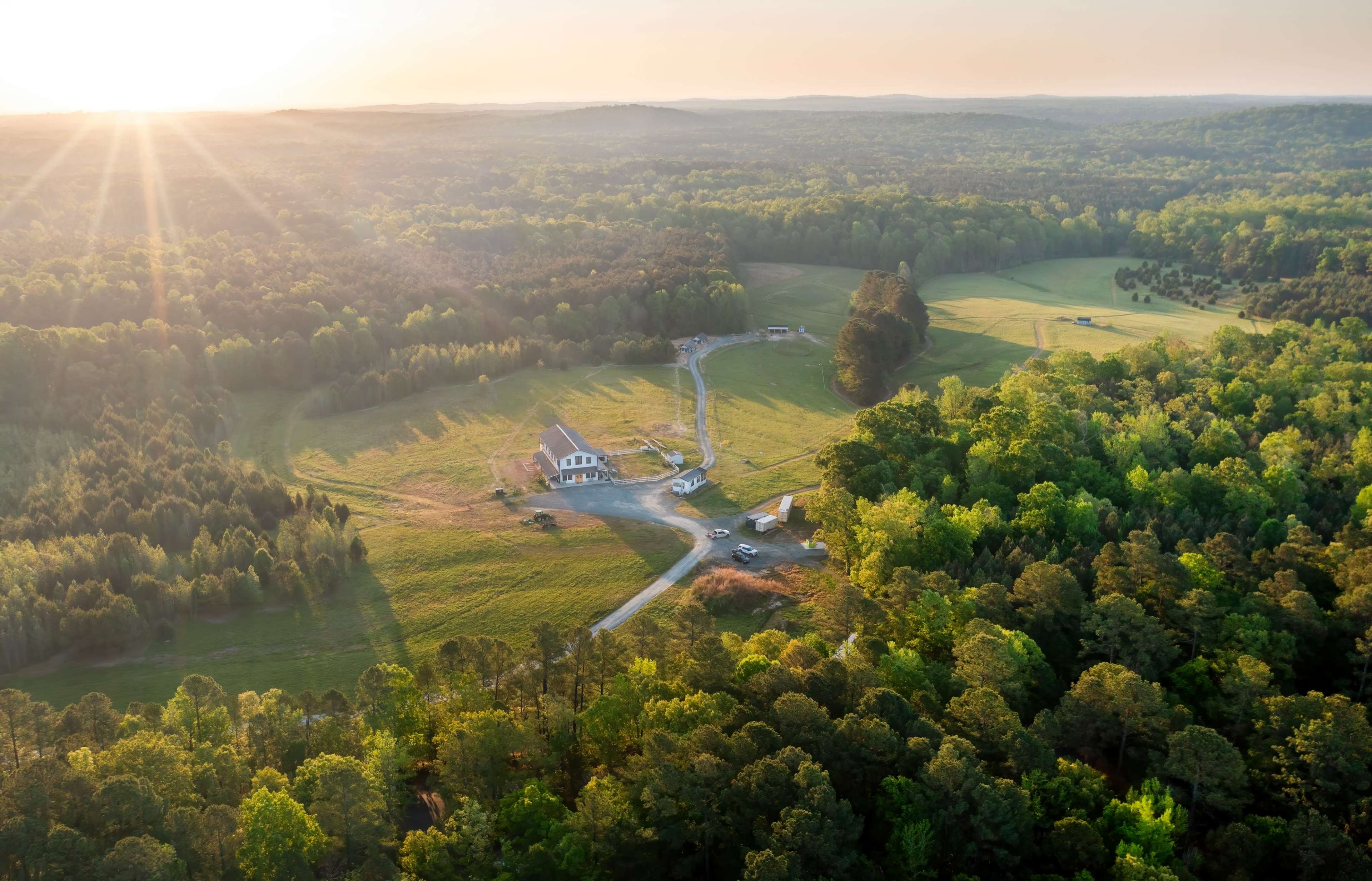 The image shows a large, green landscape with a winding road leading to a single house, surrounded by trees and open fields under a setting sun.