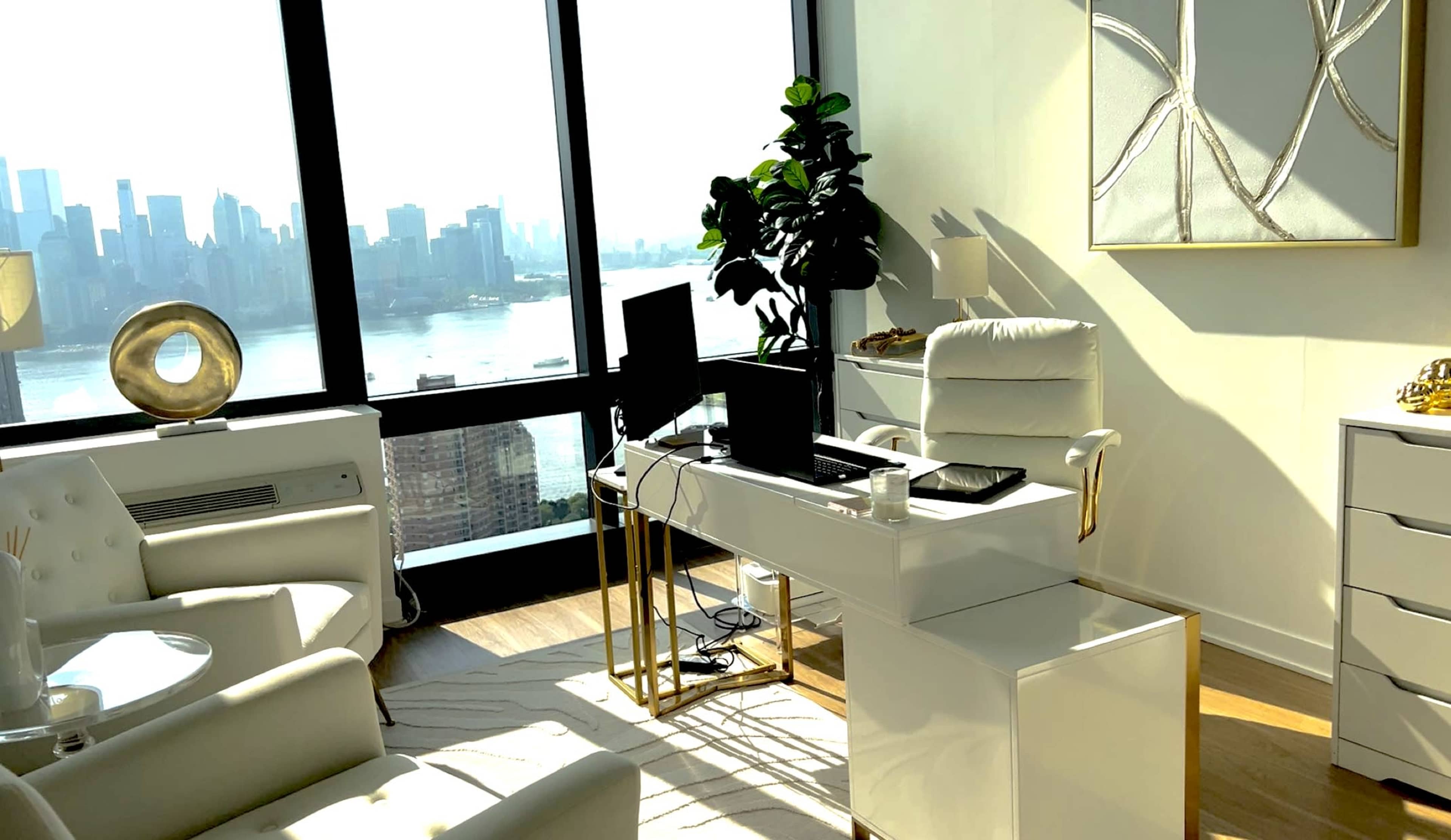 The image shows a modern office space with white furniture, a large window overlooking a city skyline, and a potted plant in the corner.
