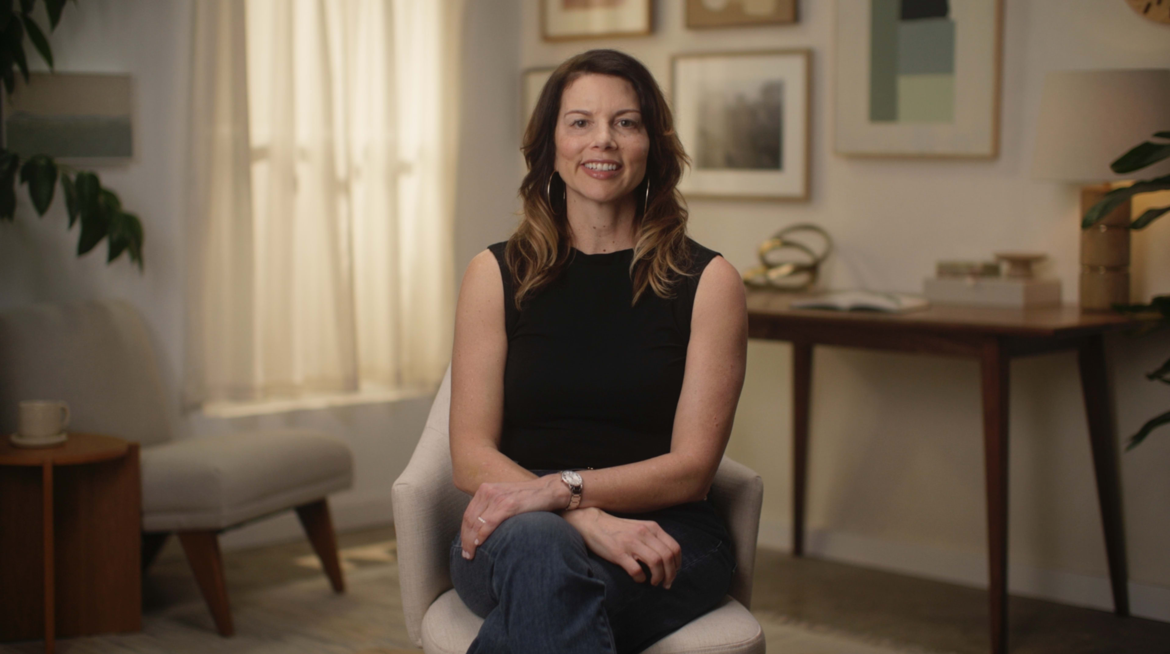 A woman sits in a chair, smiling, in a well-lit room adorned with framed artwork and a wooden table.