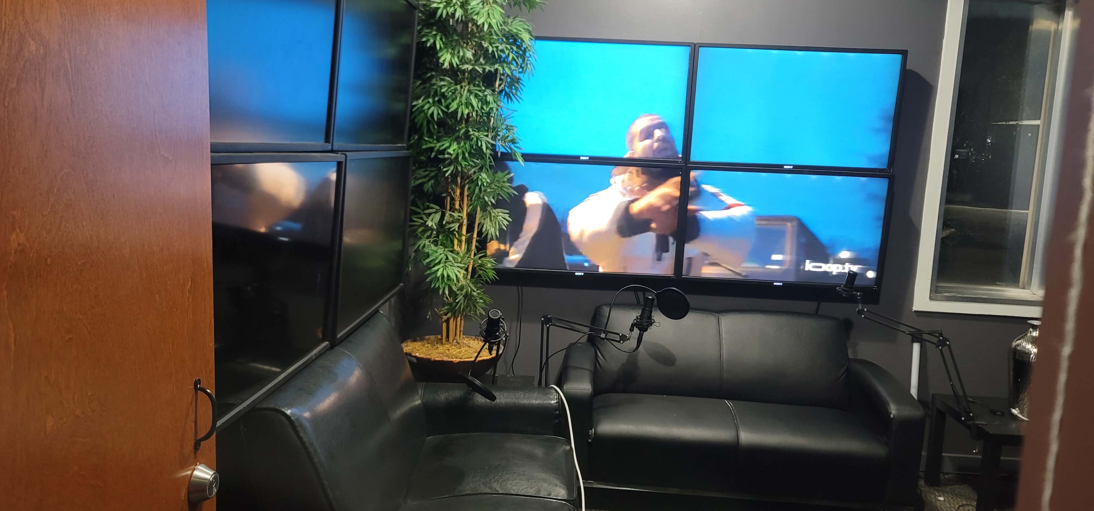 A small room with black leather couches, a large multi-screen display on the wall, and a potted plant in the corner.