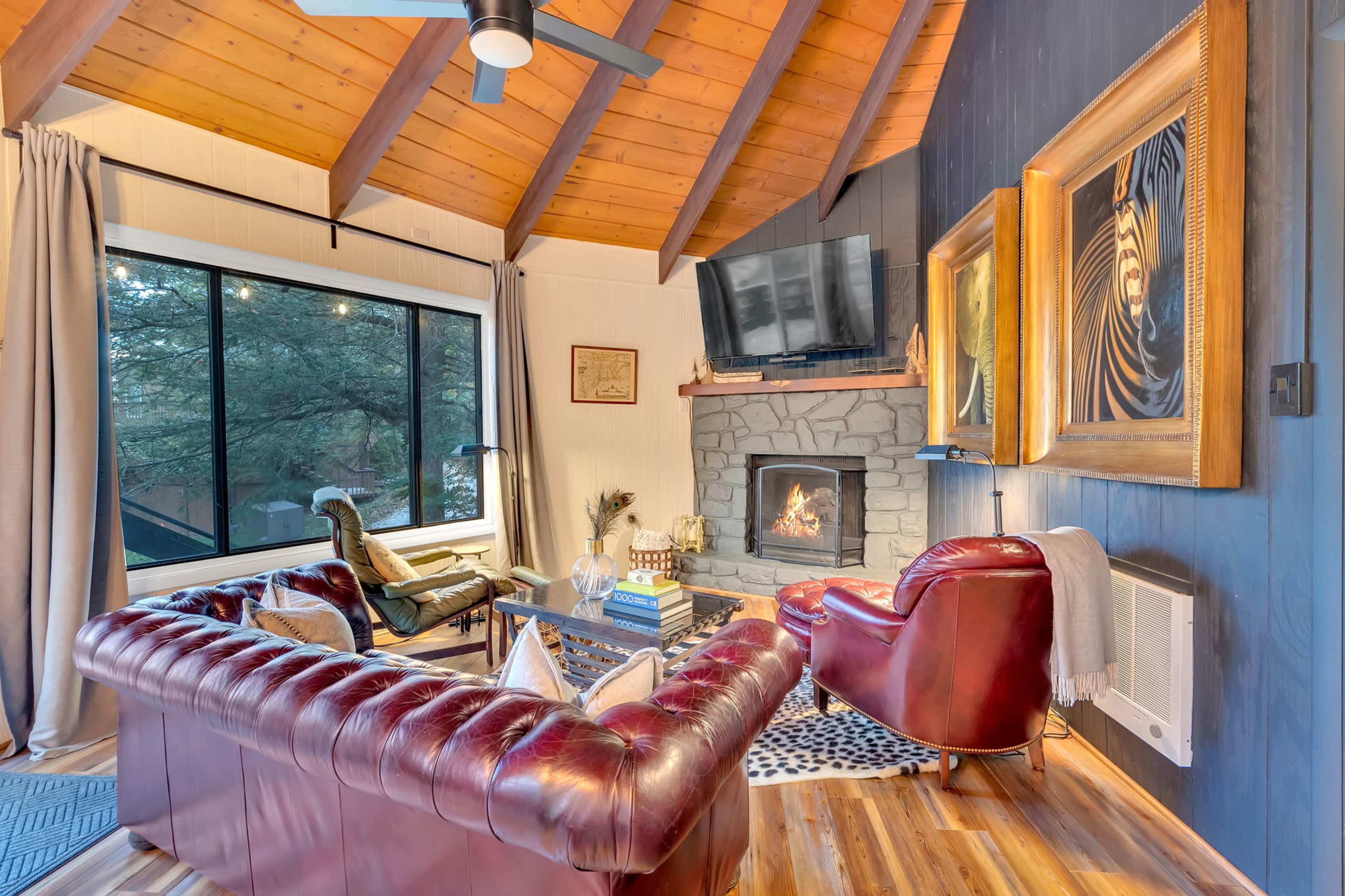 A cozy living room with a stone fireplace, a large window, and leather furniture under a wooden beam ceiling.