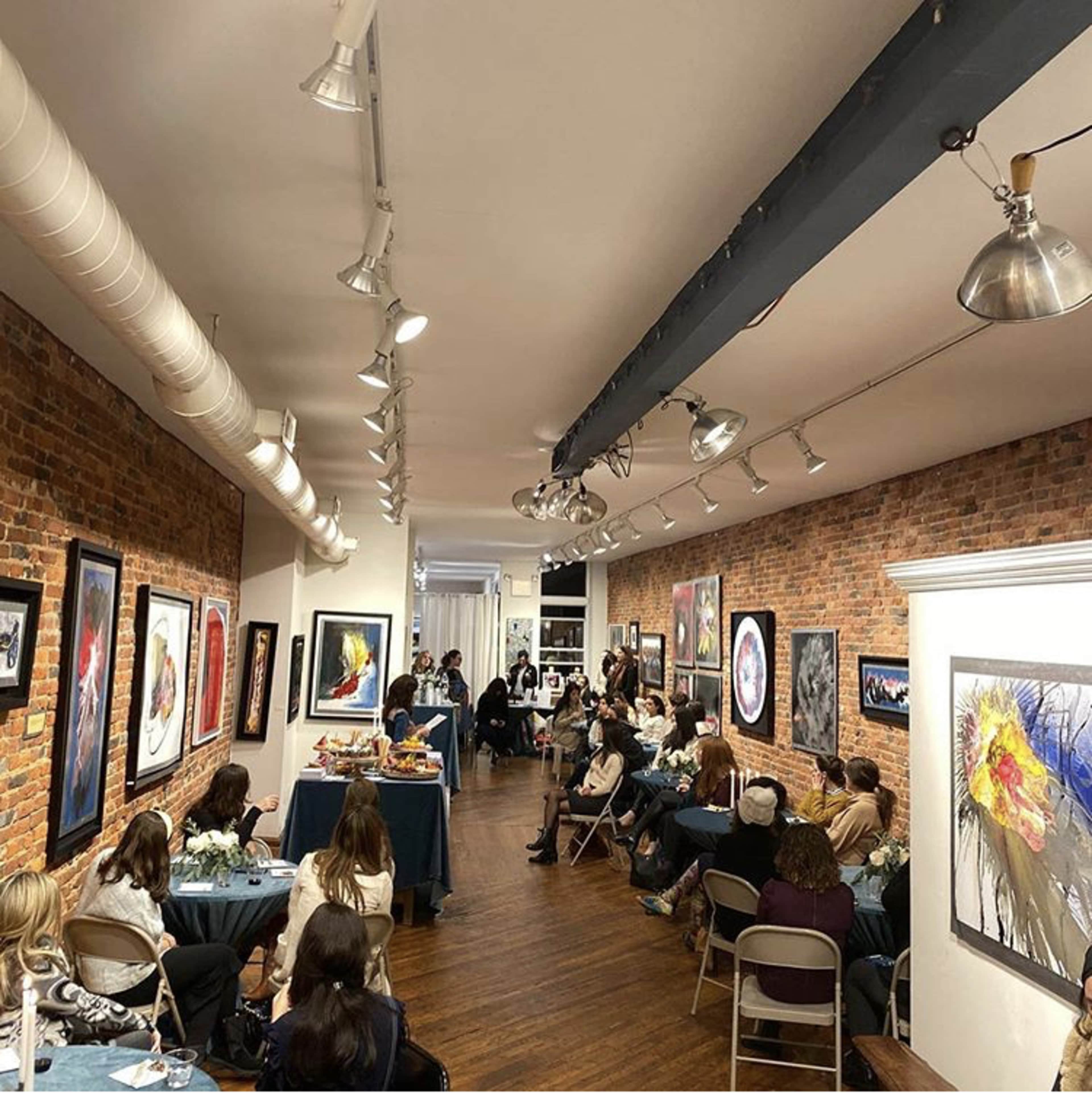 A group of people sits in a gallery with brick walls, surrounded by various framed artworks, while an event takes place at the front of the room.