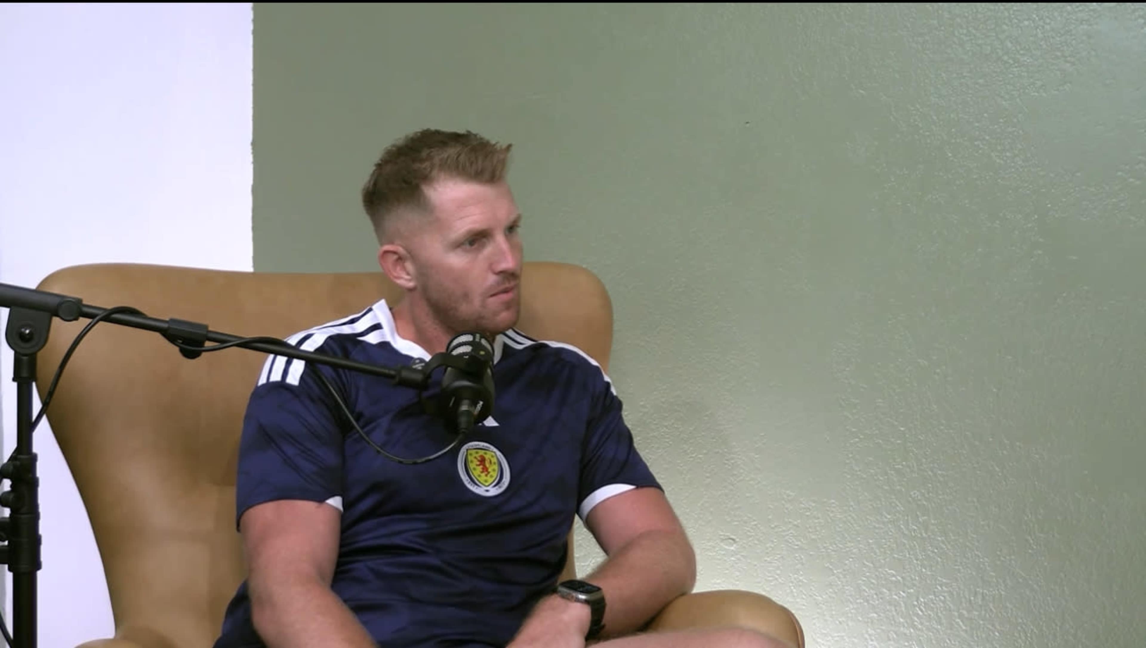 A man wearing a blue Scotland football jersey sits in a brown chair with a microphone in front of him.