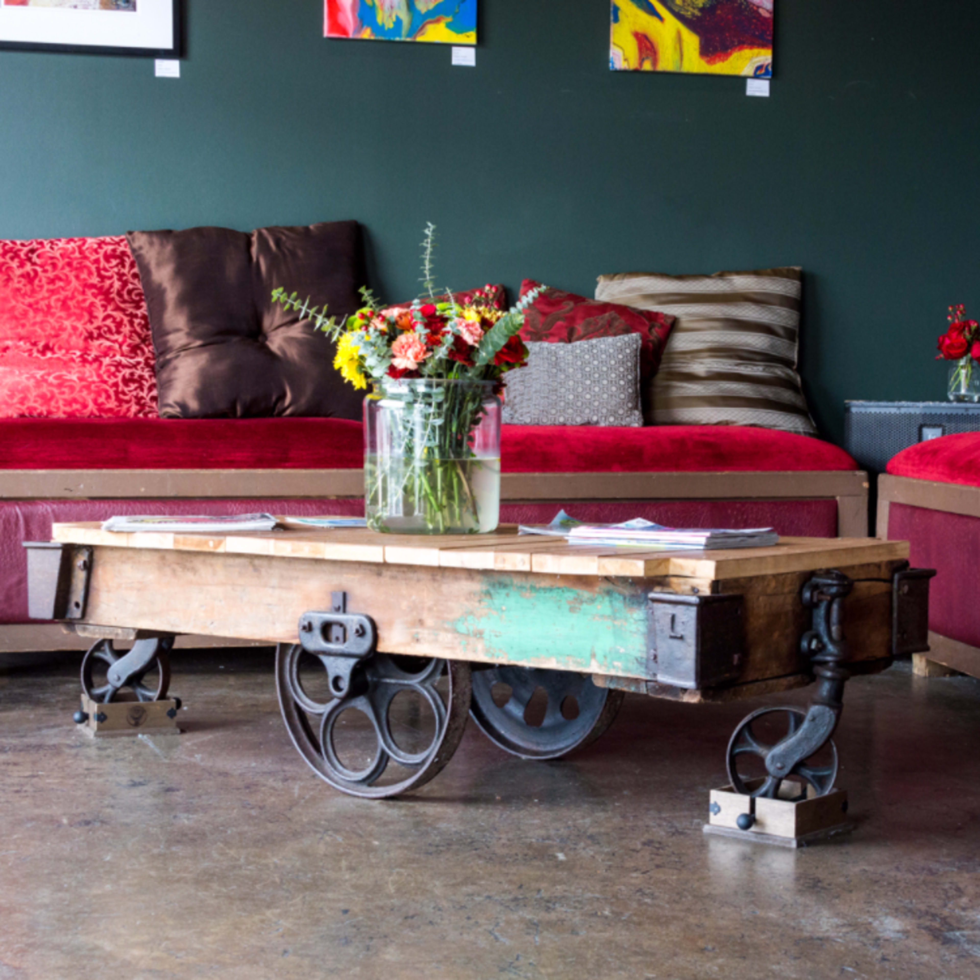A wooden coffee table on wheels is positioned in a lounge area with red sofas and colorful artwork on the walls.