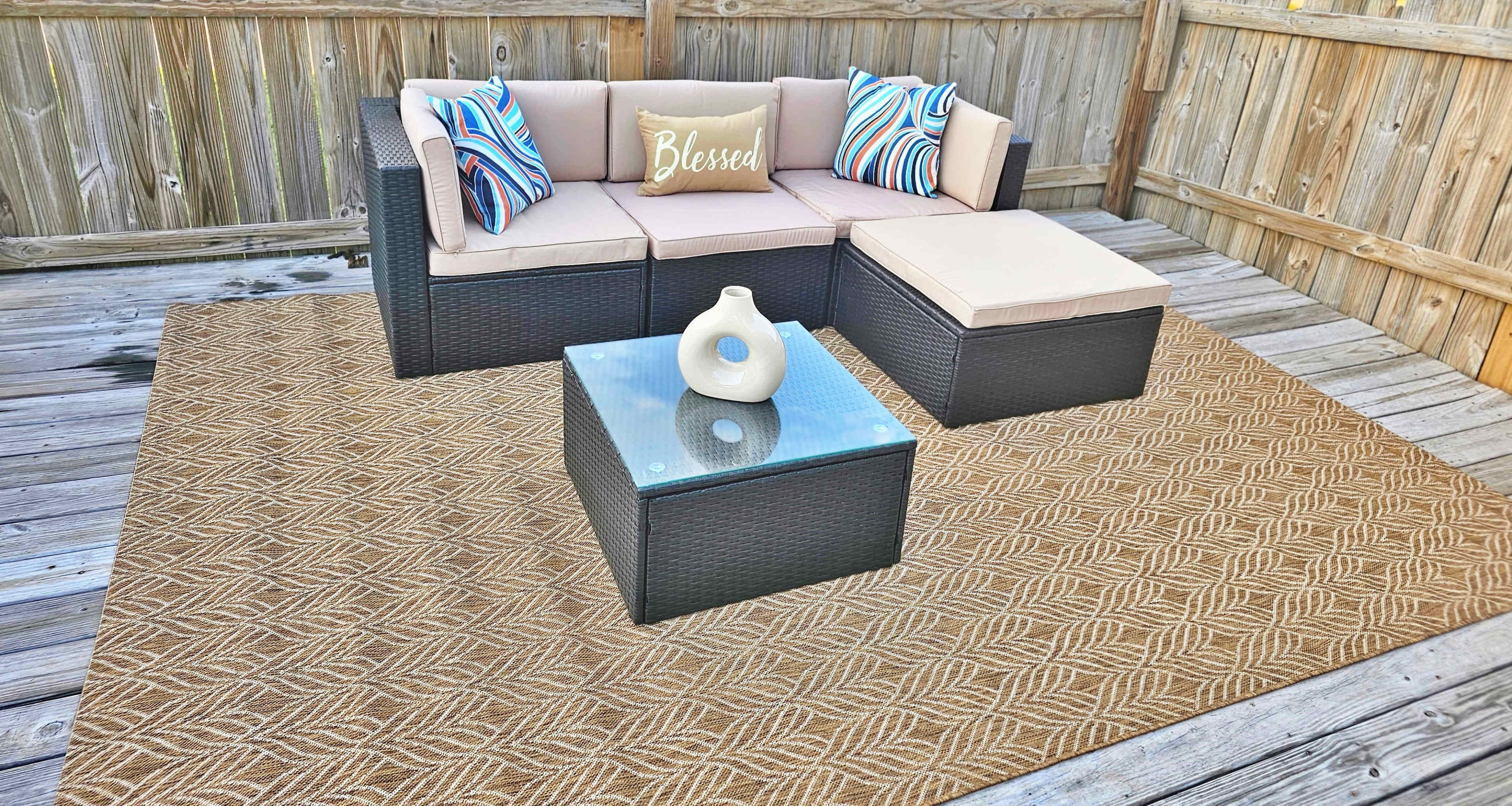 A outdoor seating area features a sectional sofa with decorative cushions, a glass-topped coffee table, and a woven rug on a wooden deck surrounded by a wooden fence.
