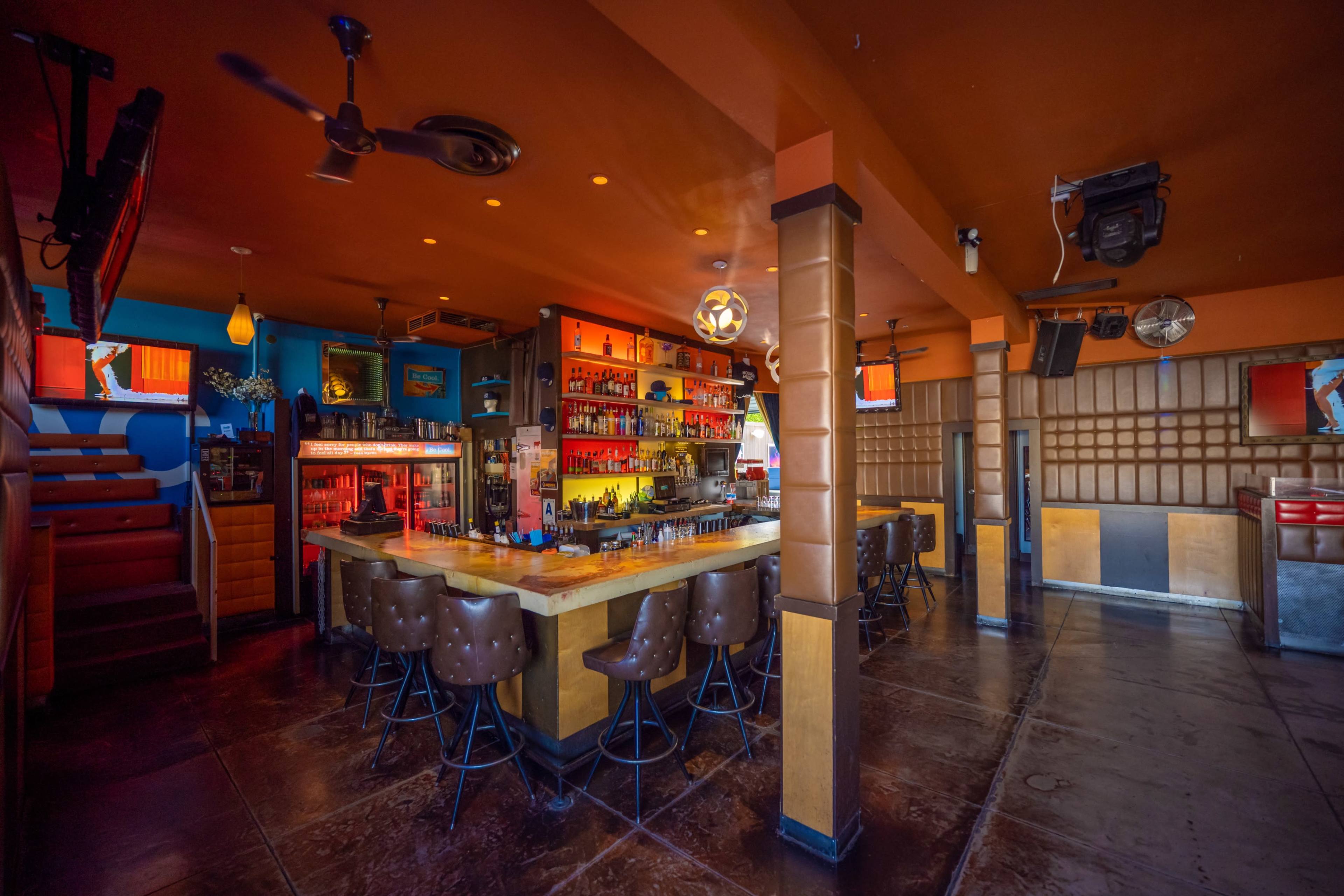 The image shows a brightly lit bar area with a well-stocked countertop and several high-backed stools, set in a vibrant, colorfully decorated interior.