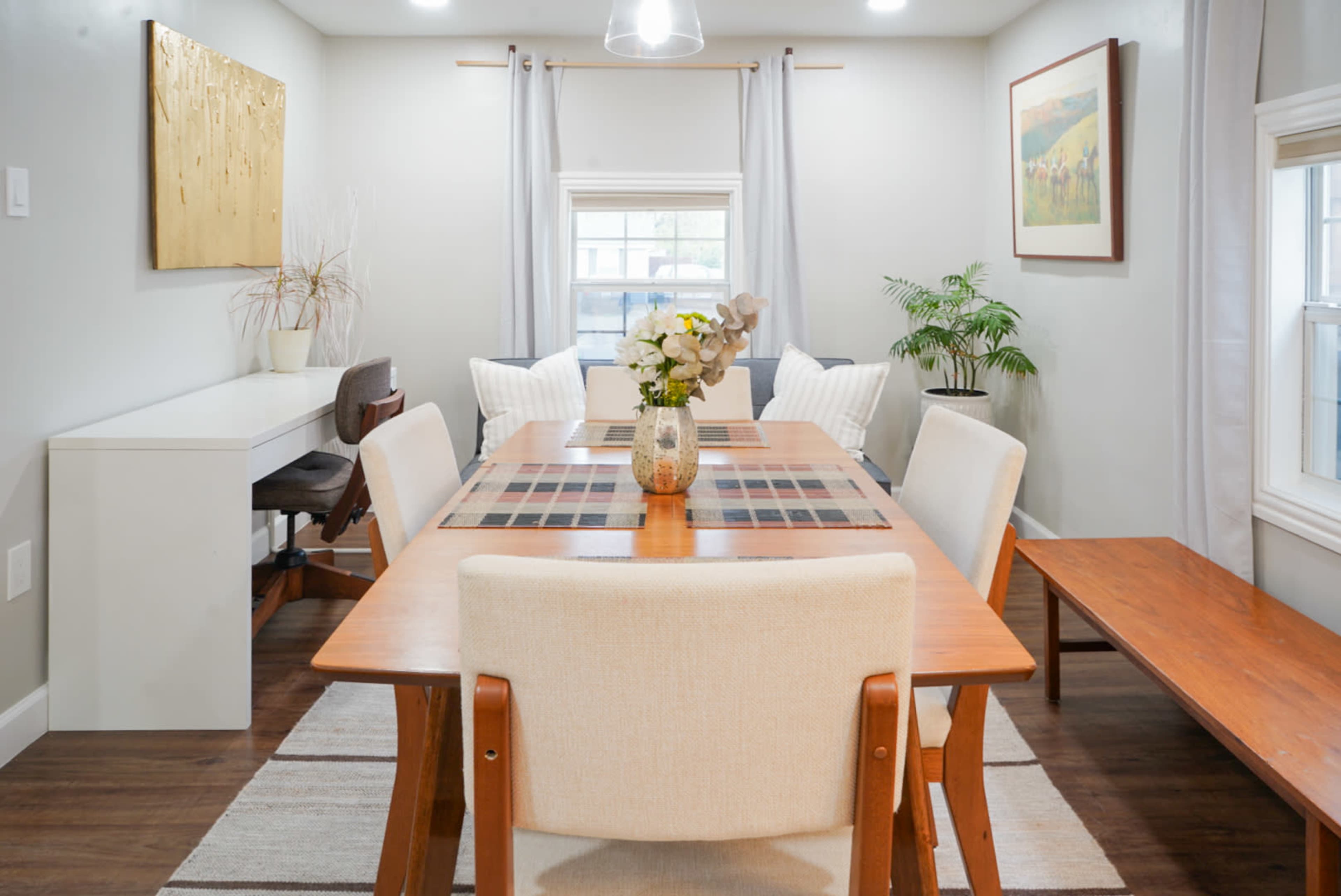 A modern dining room features a rectangular table surrounded by upholstered chairs, with a plant and artwork on the walls.