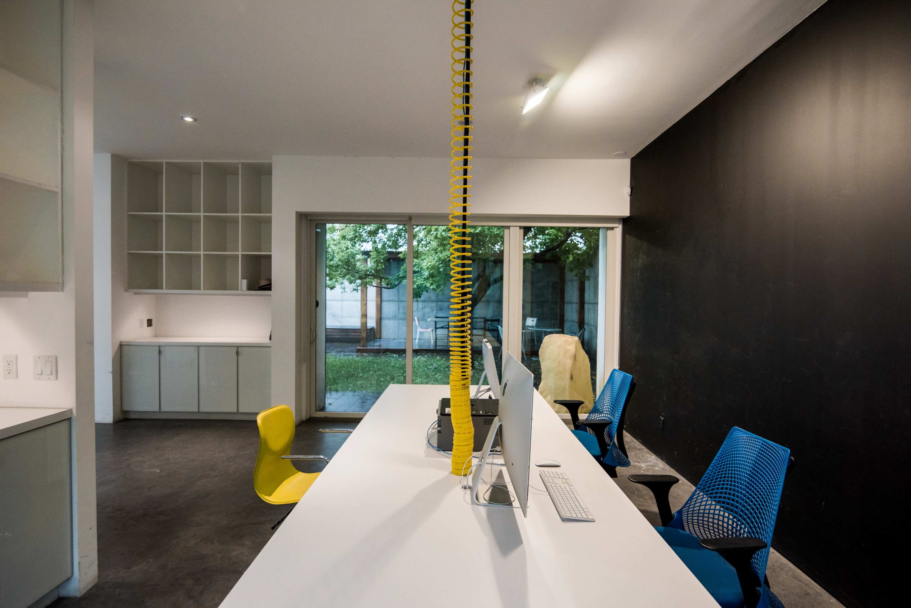 The image shows a modern office space with a long white desk, two colorful chairs, and a large window overlooking a small garden.