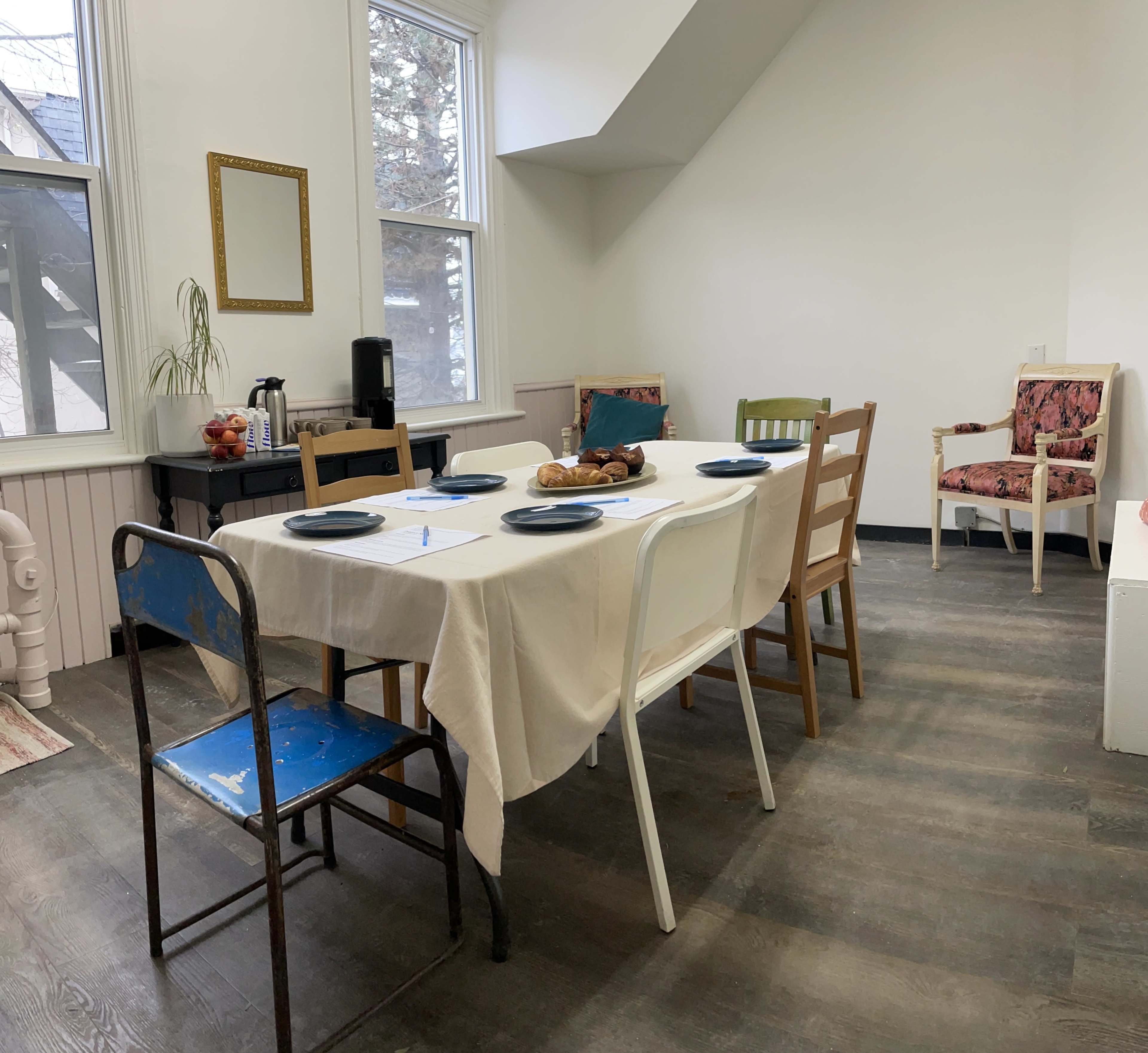 A dining area features a table set for a meal, surrounded by various chairs against a light-colored wall.