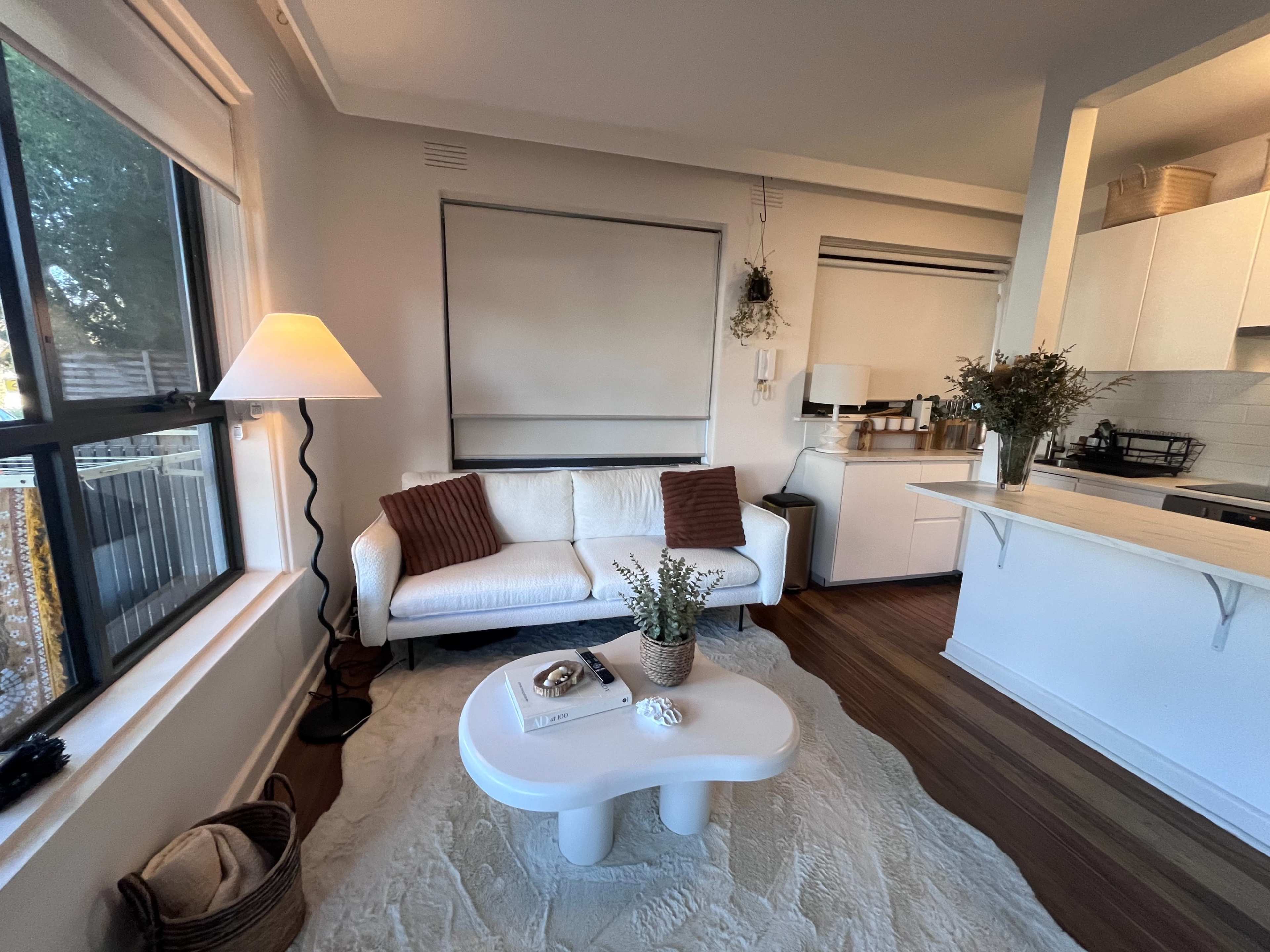 A living room with a white couch, a round coffee table, a floor lamp, and a kitchen area in the background.