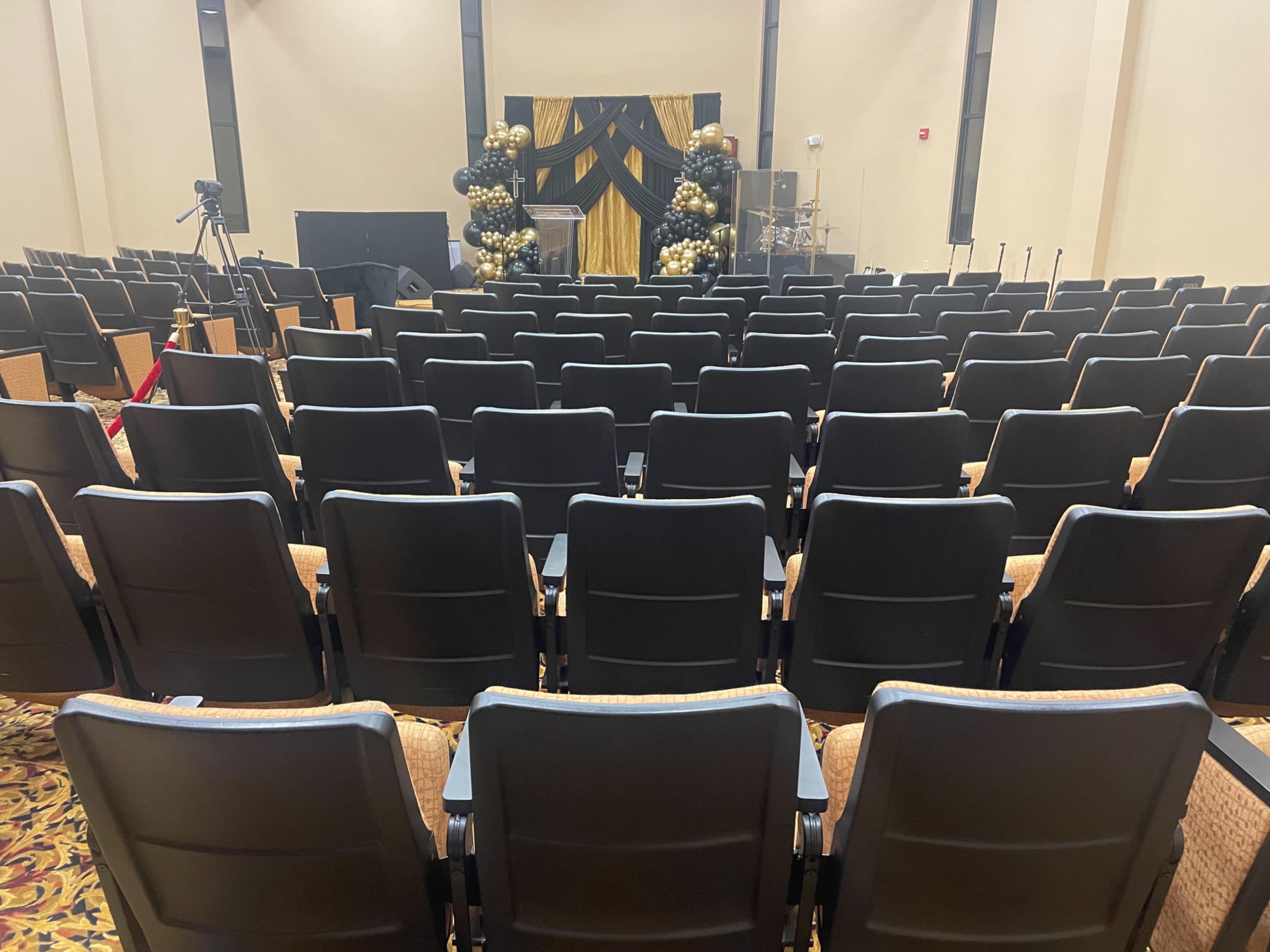 The image shows a nearly empty auditorium with rows of black chairs facing a stage decorated with black and gold balloons.