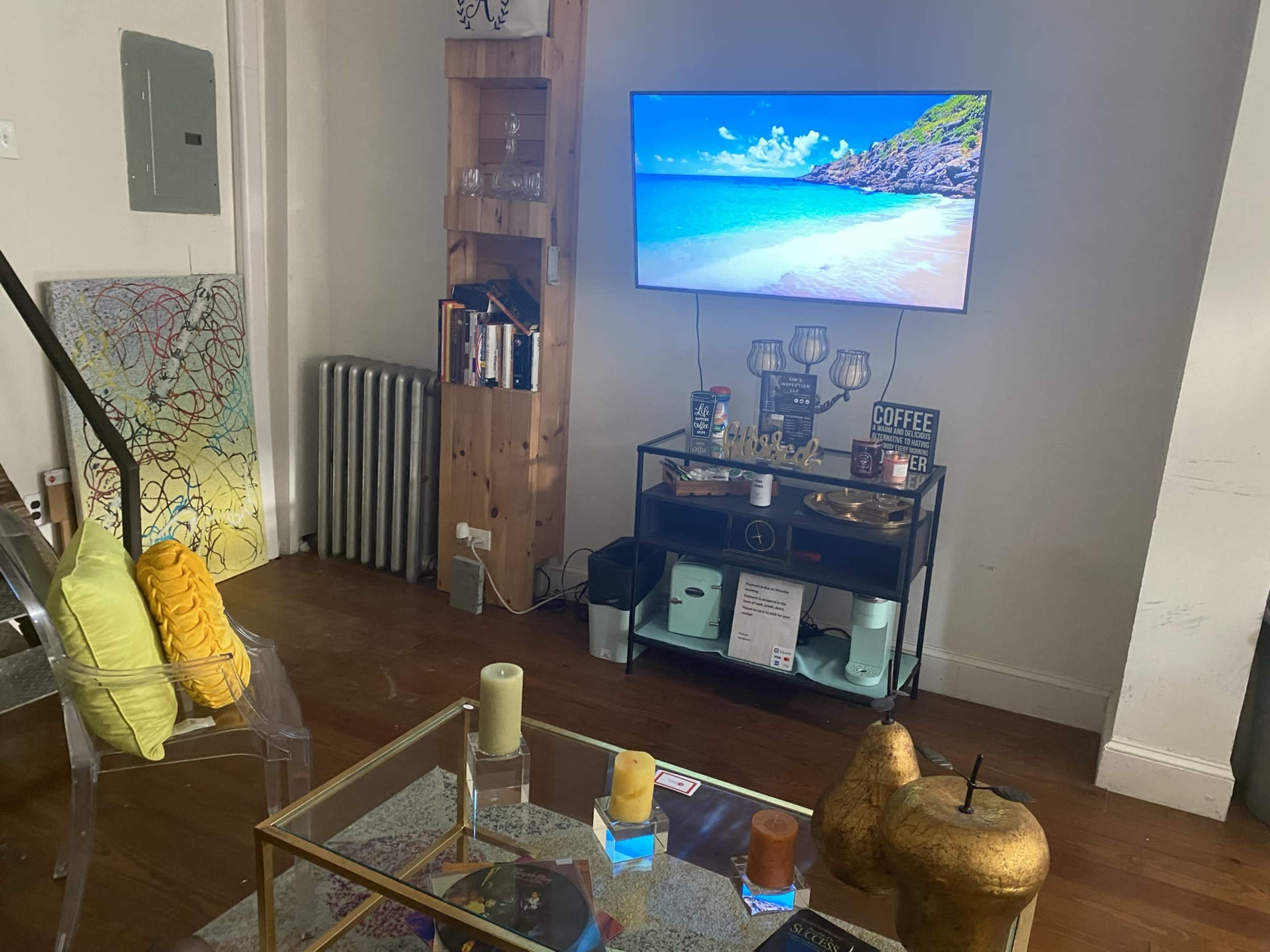 A living room features a TV mounted on the wall displaying a beach scene, with a coffee table in front, decorative items, and a colorful painting hanging nearby.