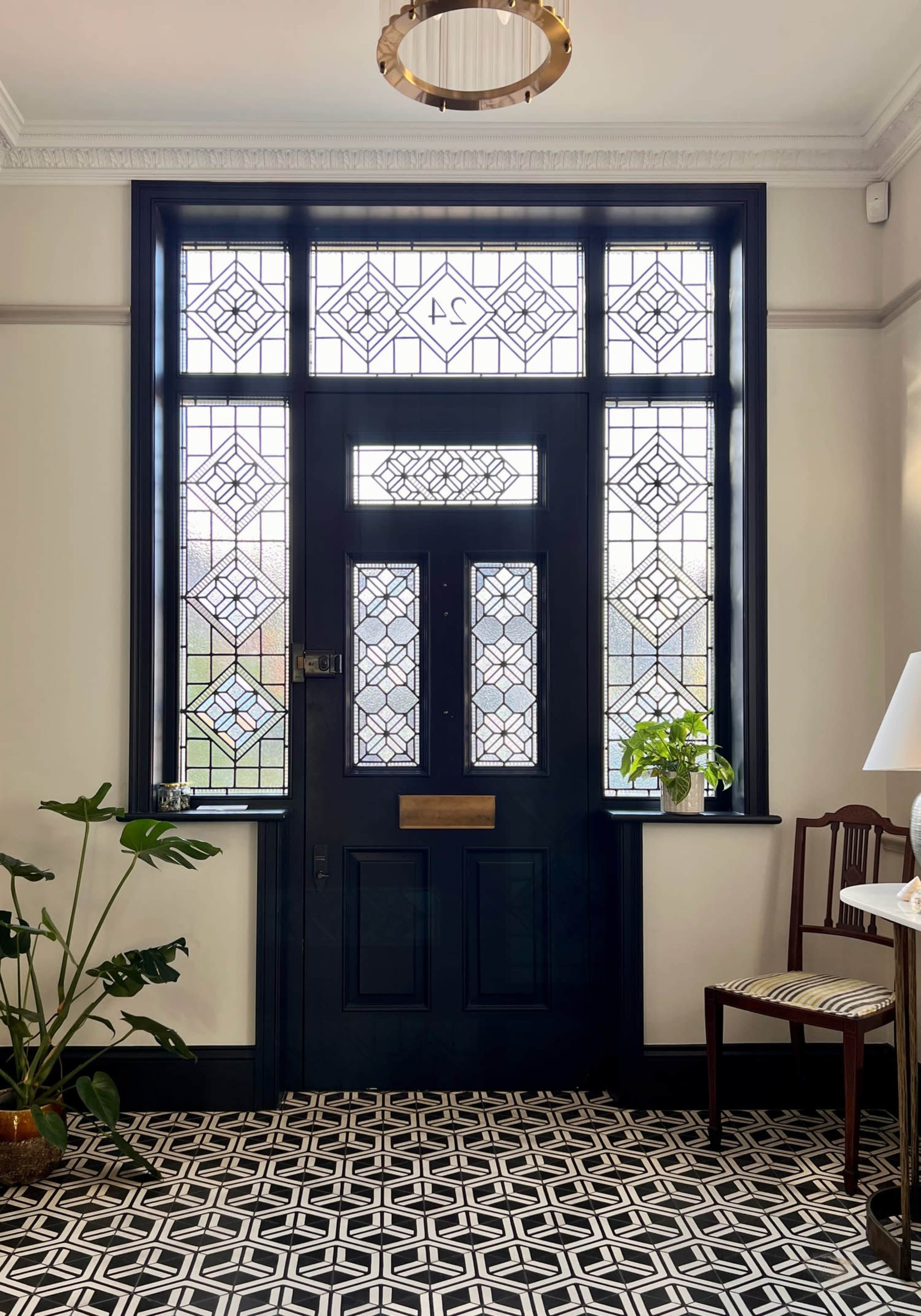 An entrance hall with a black front door featuring decorative glass panels, flanked by large windows and a geometric patterned floor.