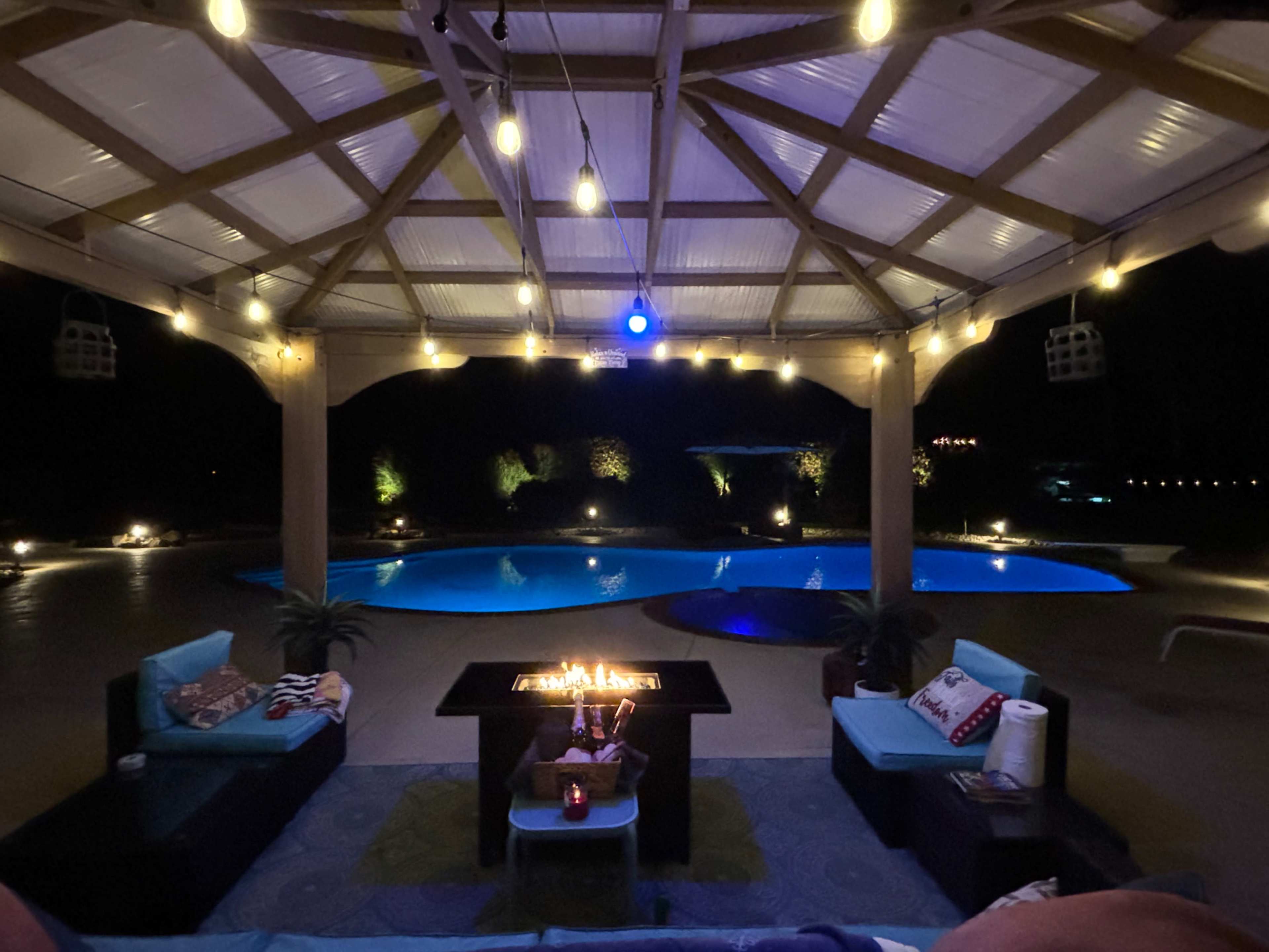 The image shows a lit outdoor pavilion overlooking a pool at night, with a fire pit surrounded by seating.