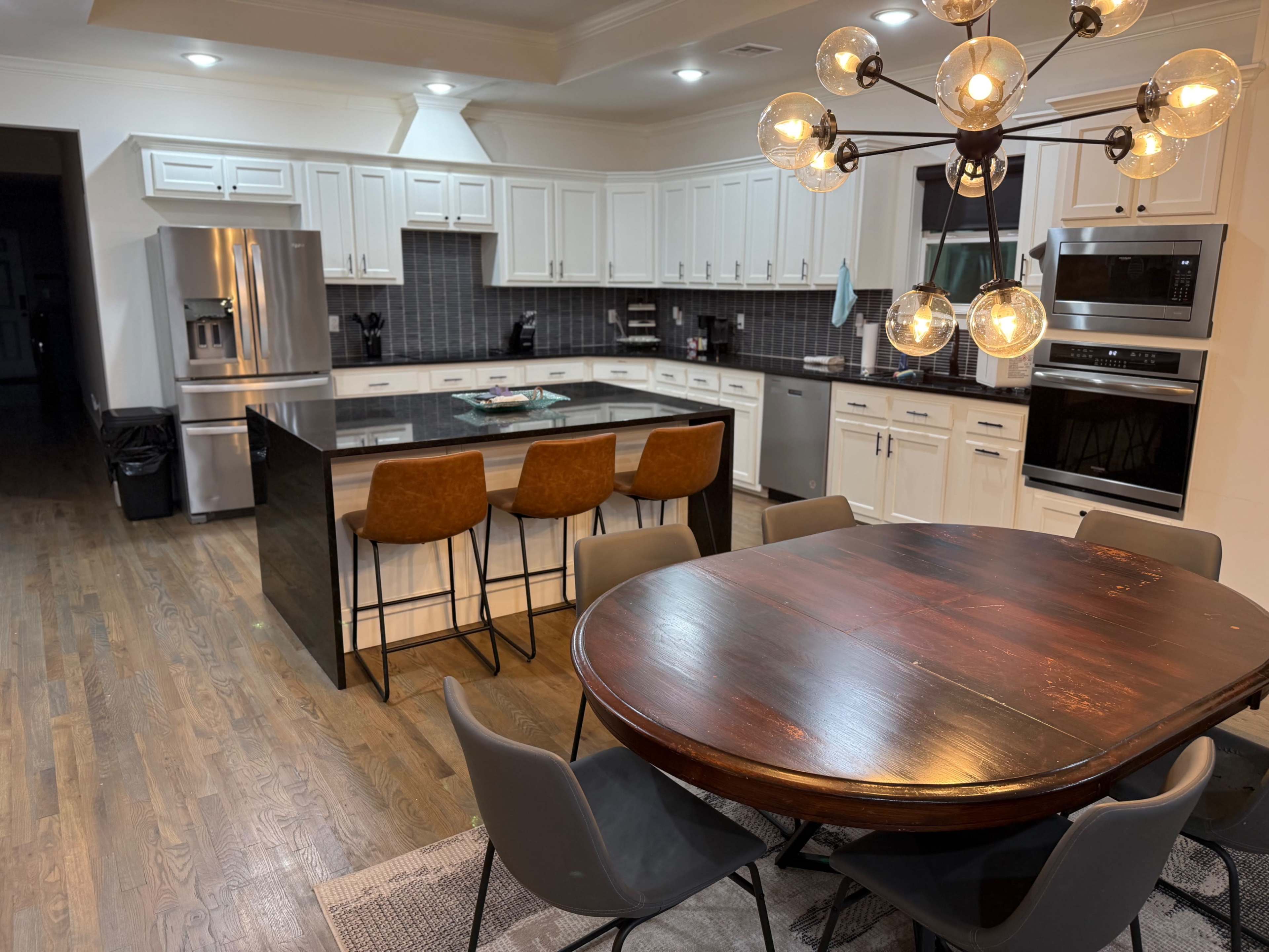 The image shows a modern kitchen with white cabinetry, a large central island with bar stools, and a dining area featuring a round wooden table surrounded by gray chairs.