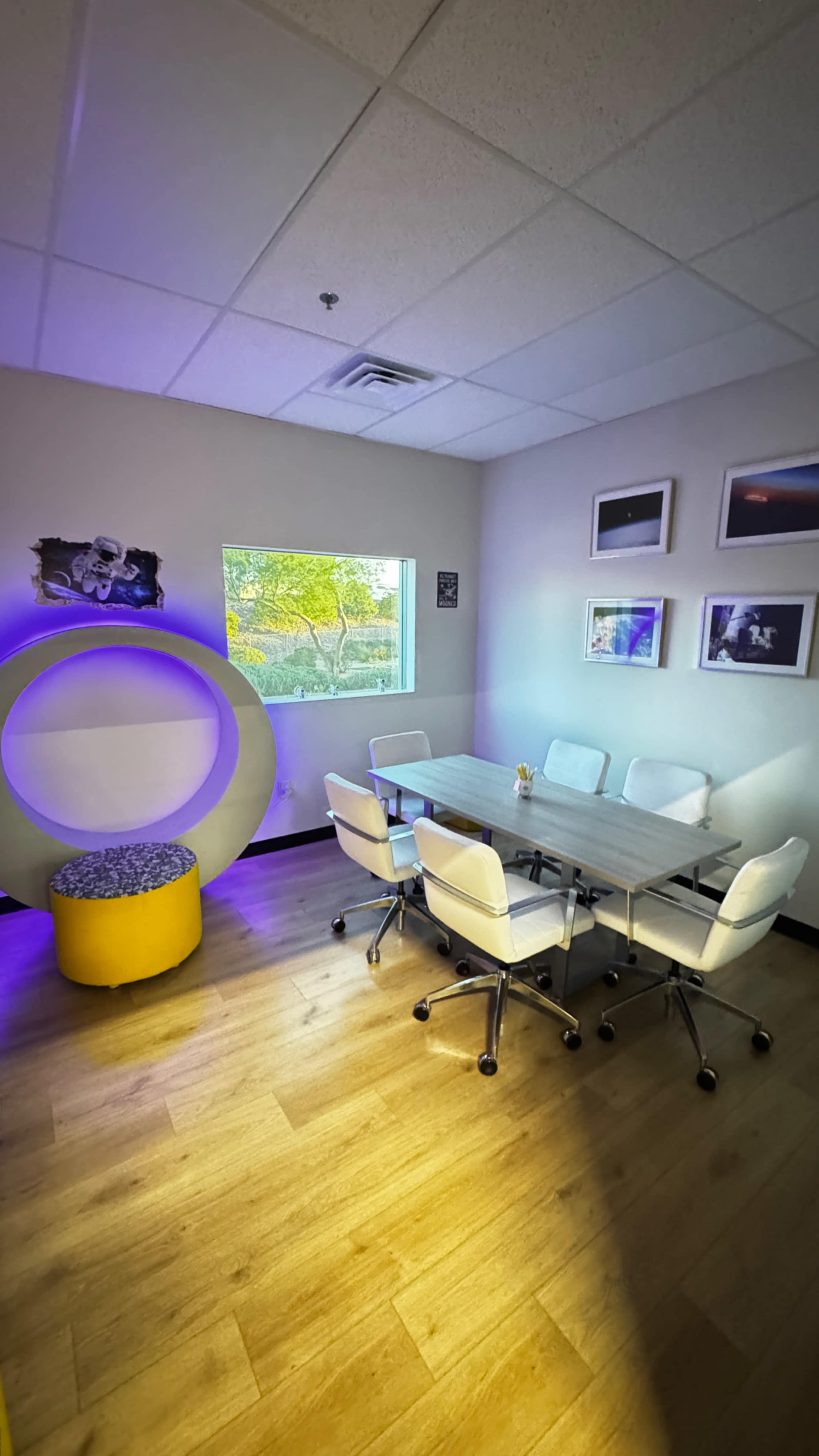 The image shows a modern conference room with a round, illuminated seating area, a wooden table surrounded by white chairs, and large windows with natural light.