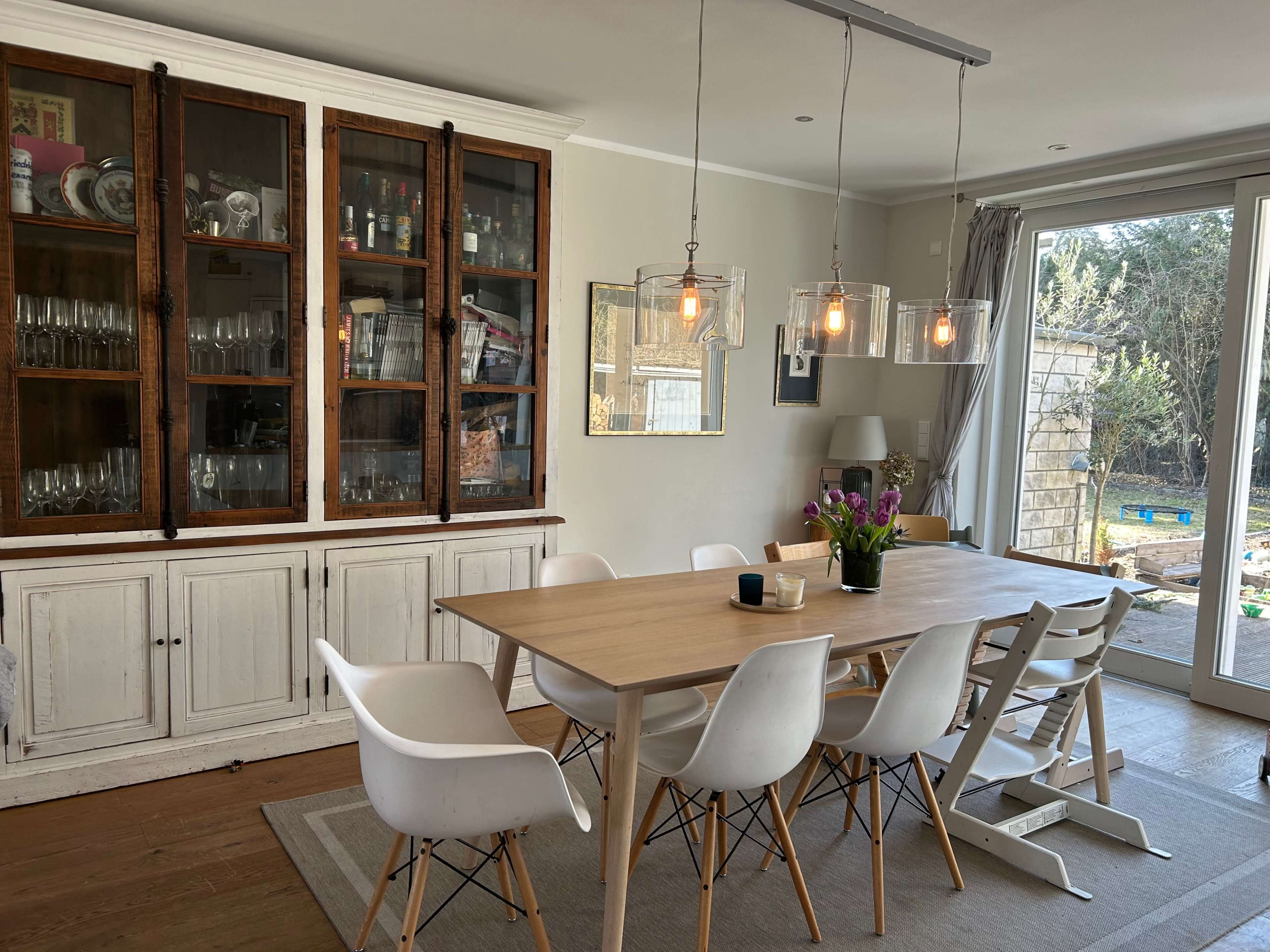 The image shows a dining room featuring a wooden table surrounded by white chairs, with a cabinet displaying glassware and a large window opening to a garden area.