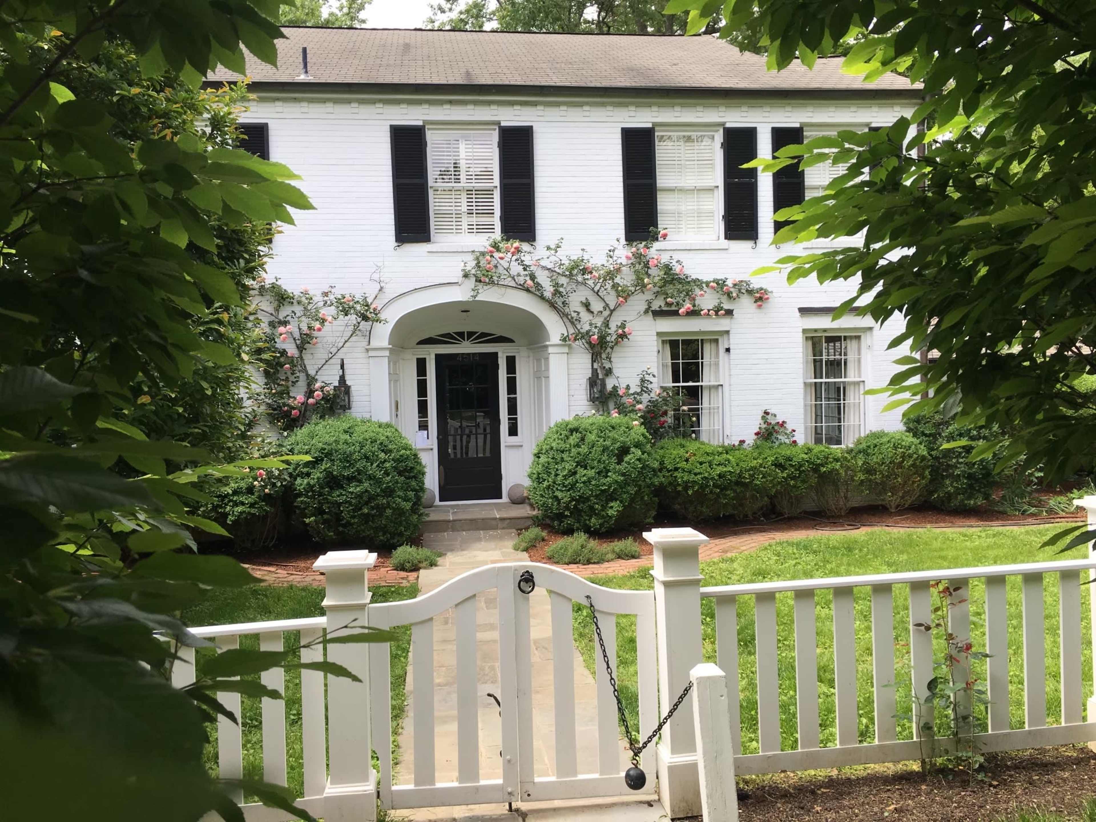 A white brick house with black shutters is framed by lush greenery and roses, featuring a covered front entrance and a white picket fence.