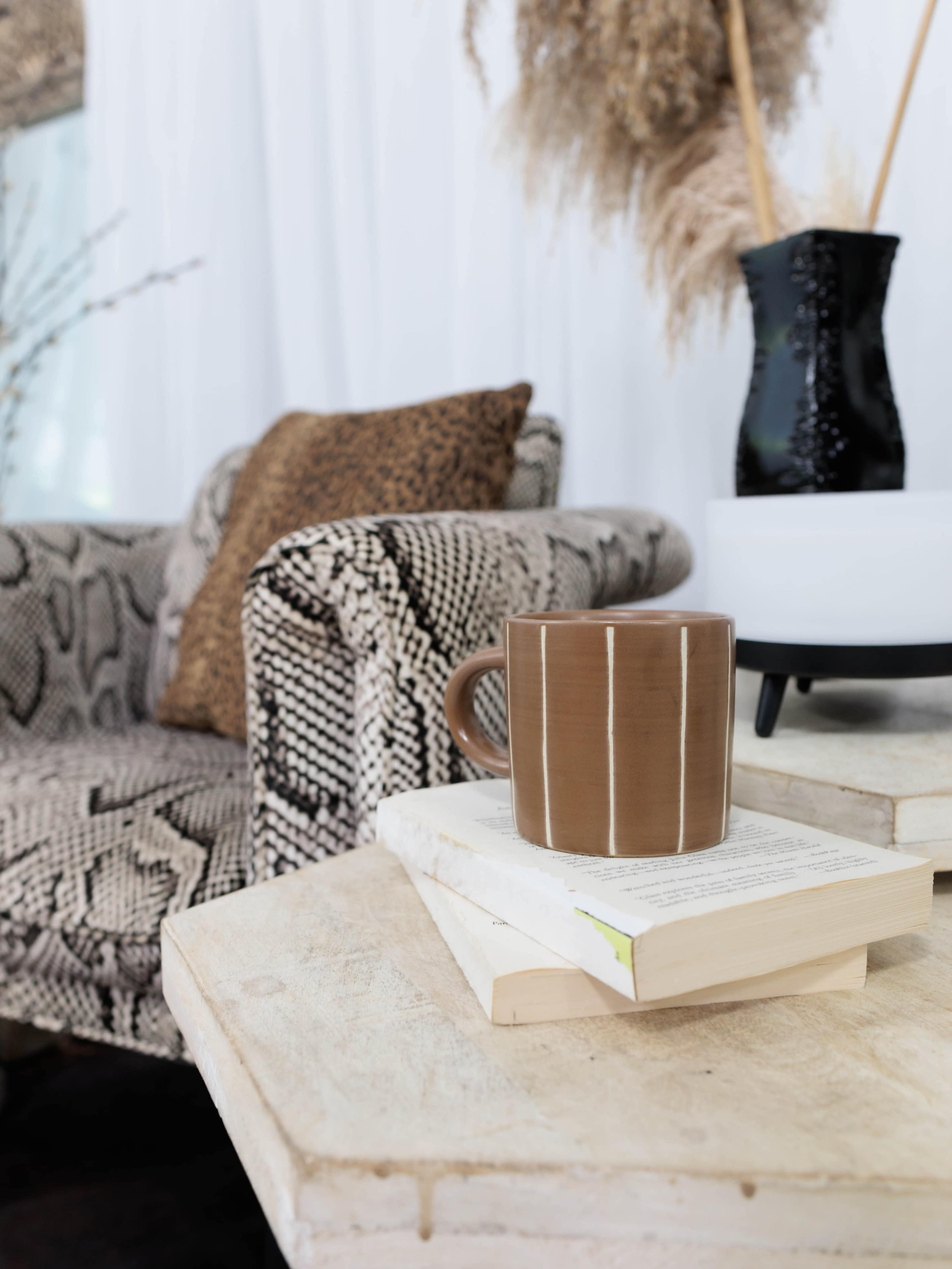 A brown mug is perched on a stack of books on a wooden table, with a patterned armchair and decorative vase in the background.