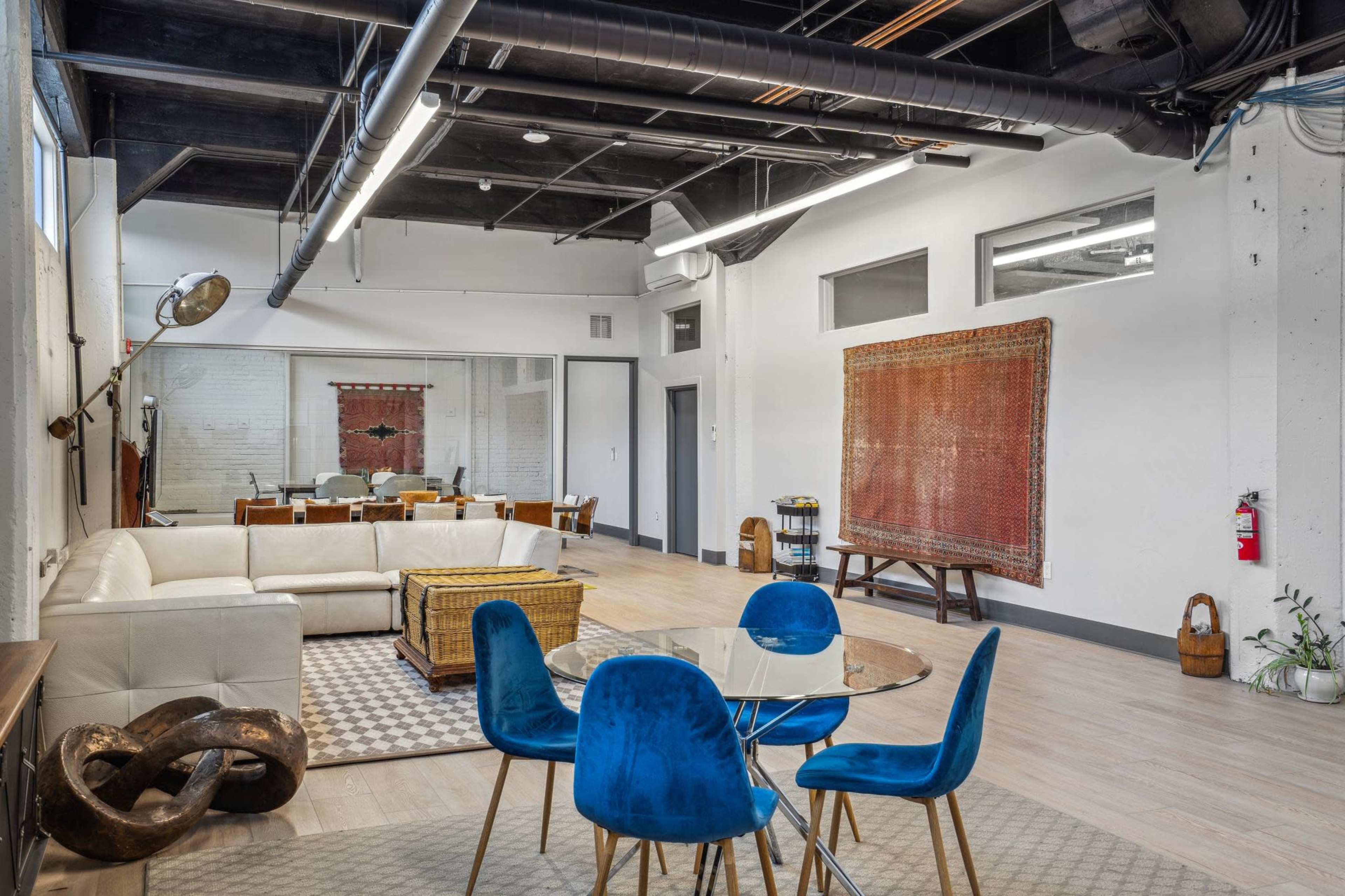 The image shows a spacious, modern loft featuring a seating area with a white couch, a dining table with blue chairs, and a large textile hanging on the wall.
