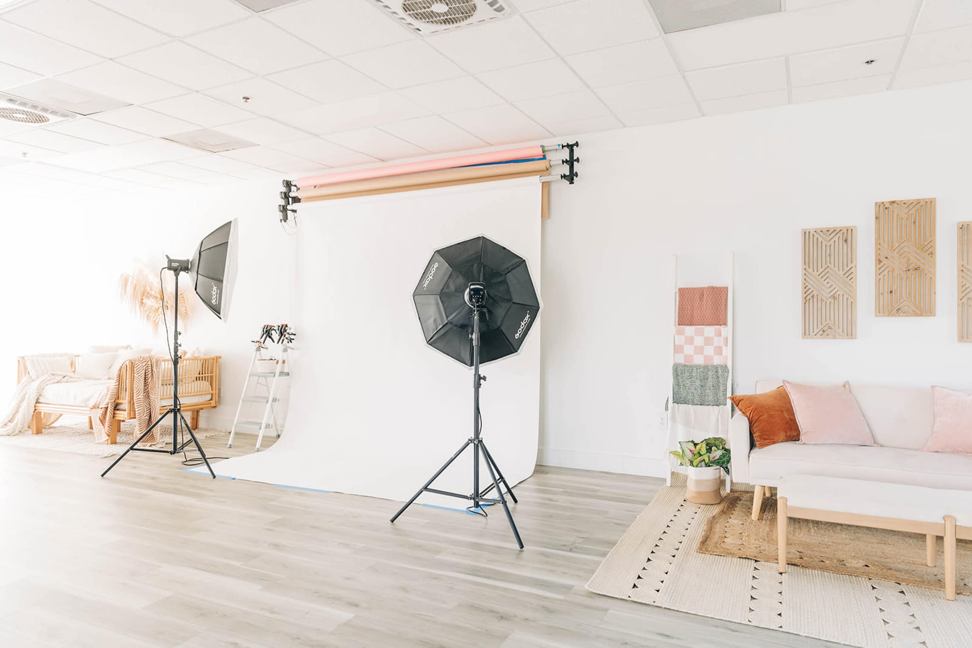 The image shows a bright photo studio setup with softbox lights, a white backdrop, a ladder, and a lounge area furnished with a sofa and decorative wall art.