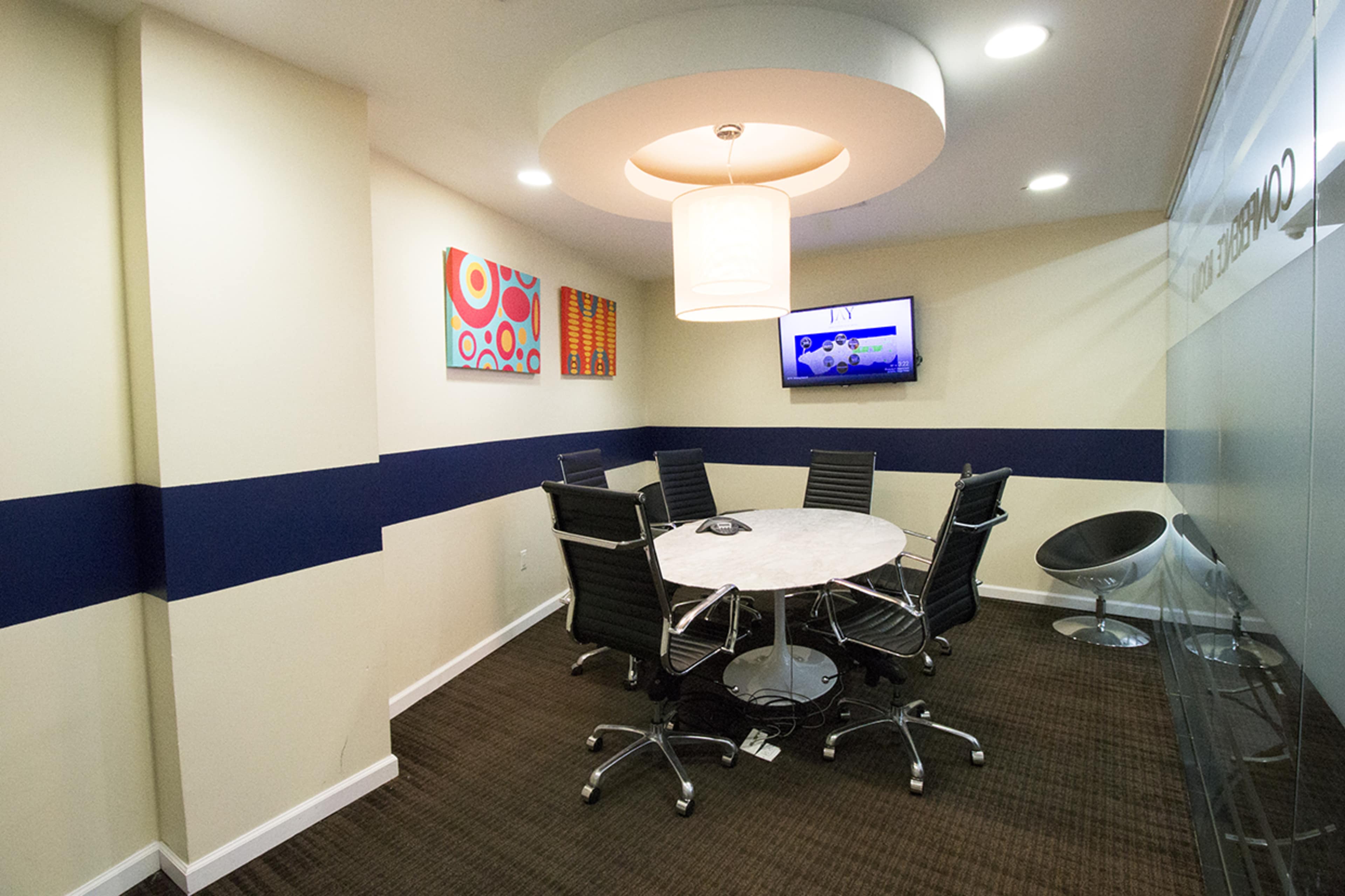 A modern meeting room with a circular table, eight chairs, a television, and wall art.