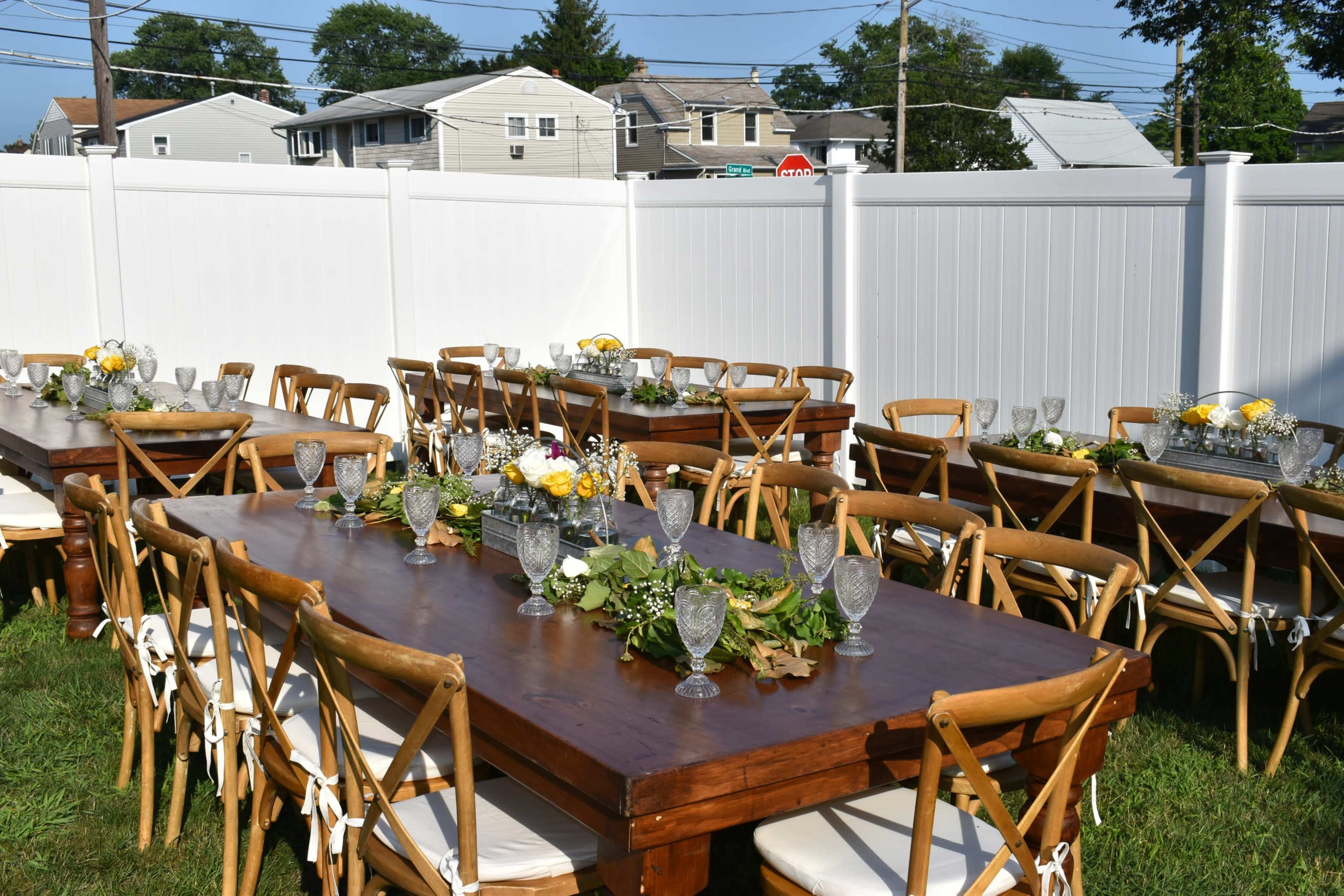 The scene features two long wooden tables set outdoors, adorned with glassware and floral centerpieces, surrounded by wooden chairs in a grassy area enclosed by a white fence.