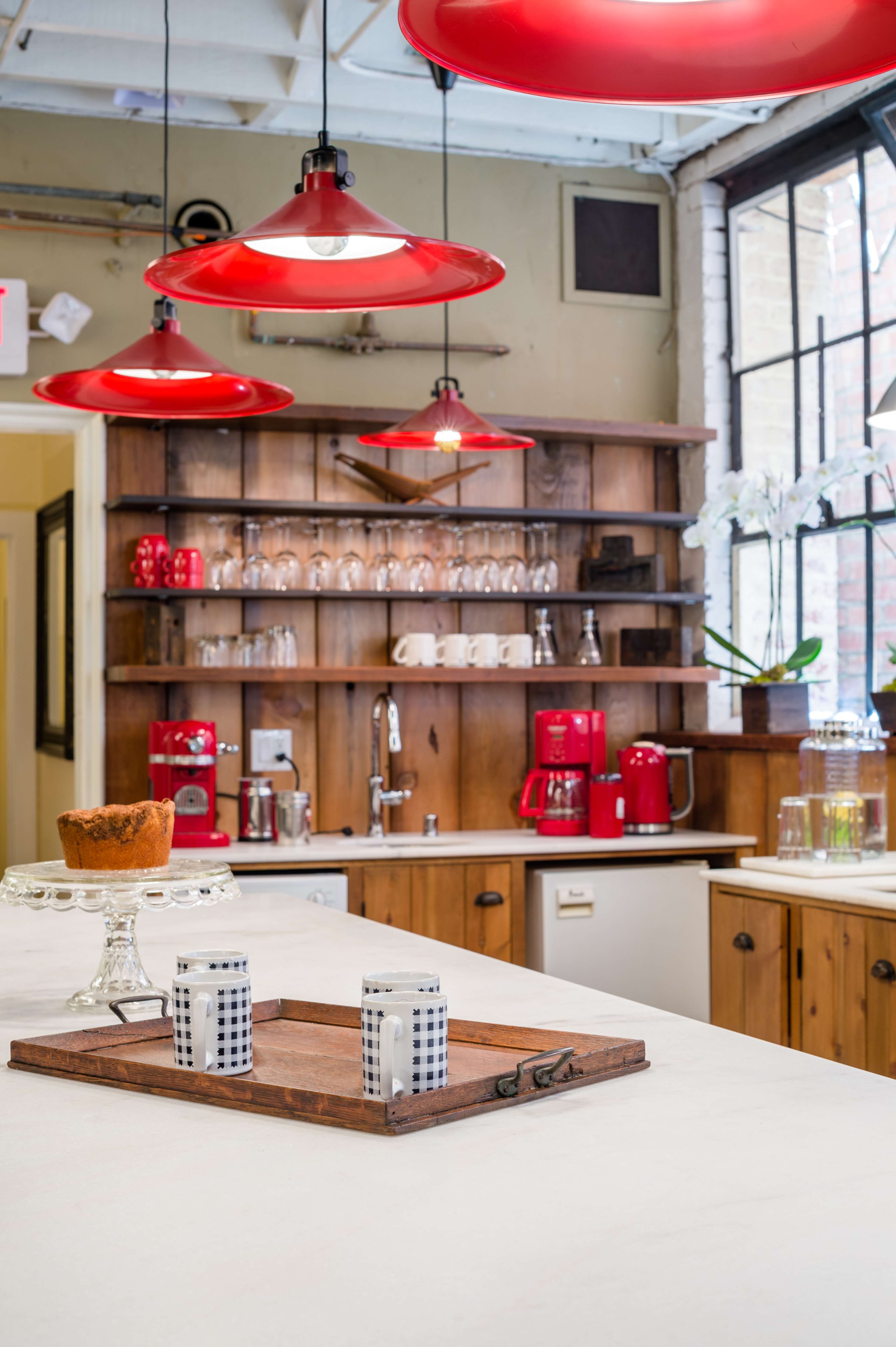 A modern kitchen features red pendant lights, wooden shelves with glassware, and a countertop display of a cake on a glass stand.