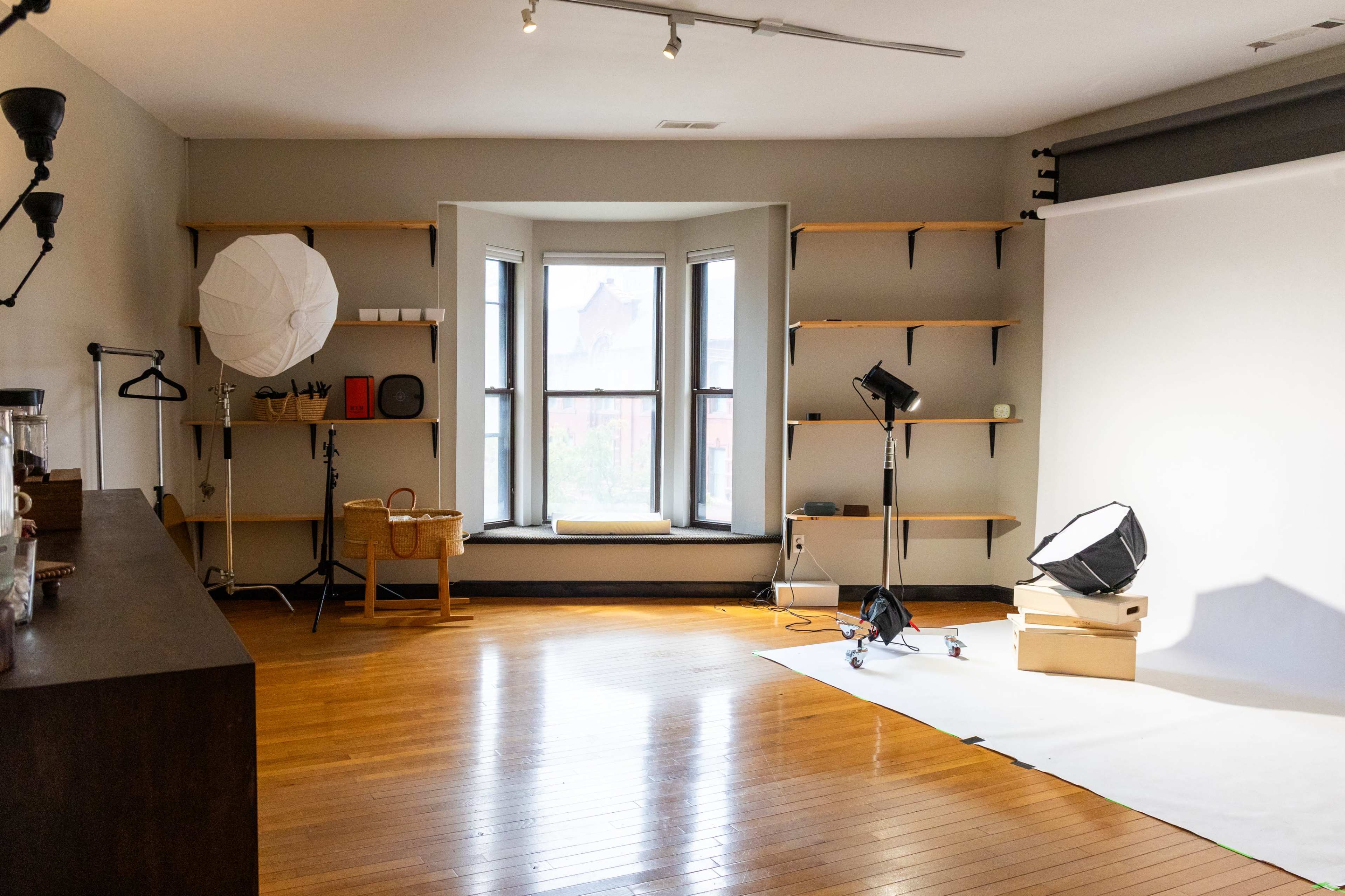 The image shows a spacious photography studio featuring a white backdrop, various lighting equipment, and wooden shelves on the walls.