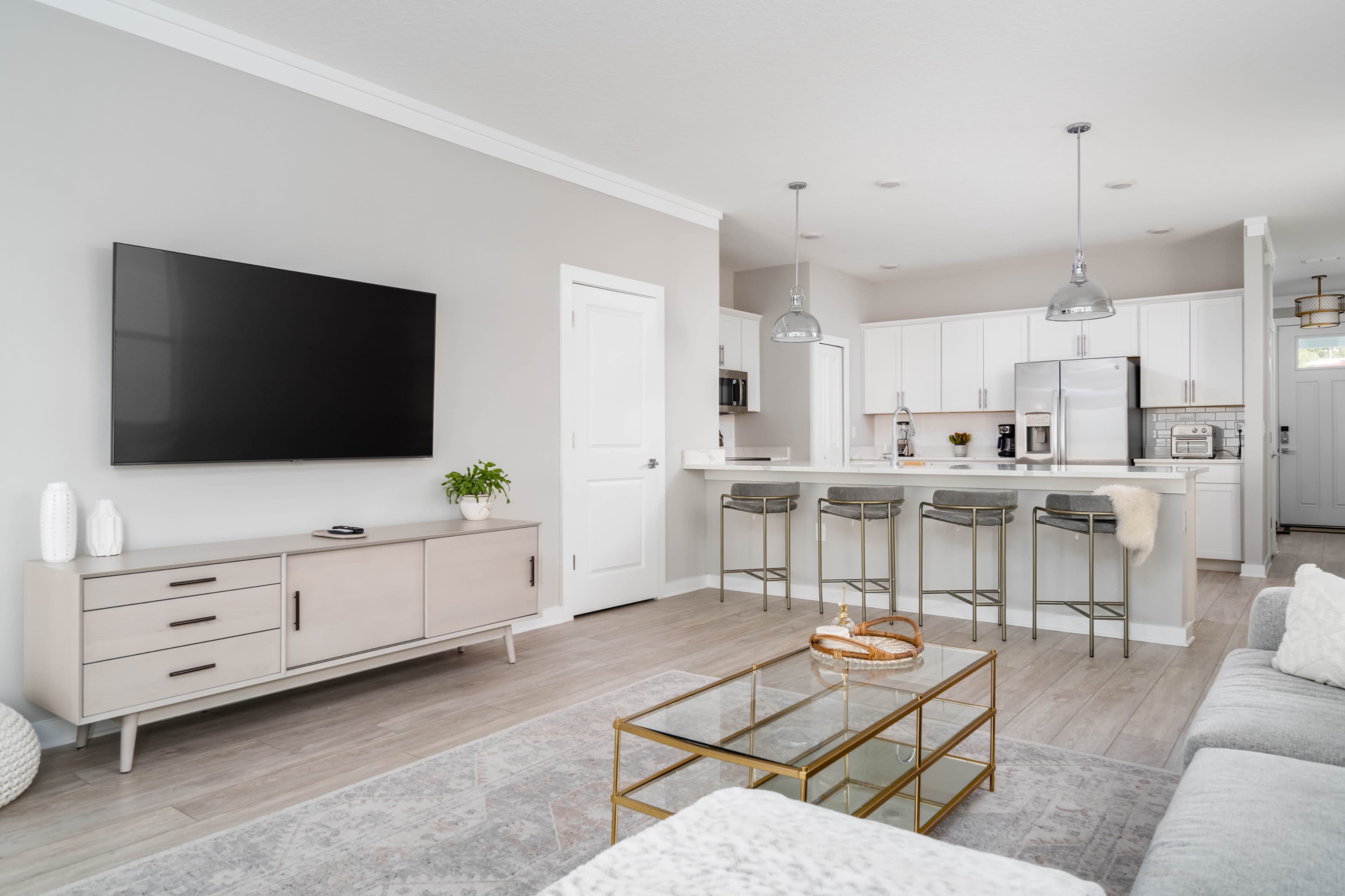 A modern living room features a flat-screen TV mounted on a light-colored wall, with a kitchen area in the background that includes white cabinets and stainless steel appliances.