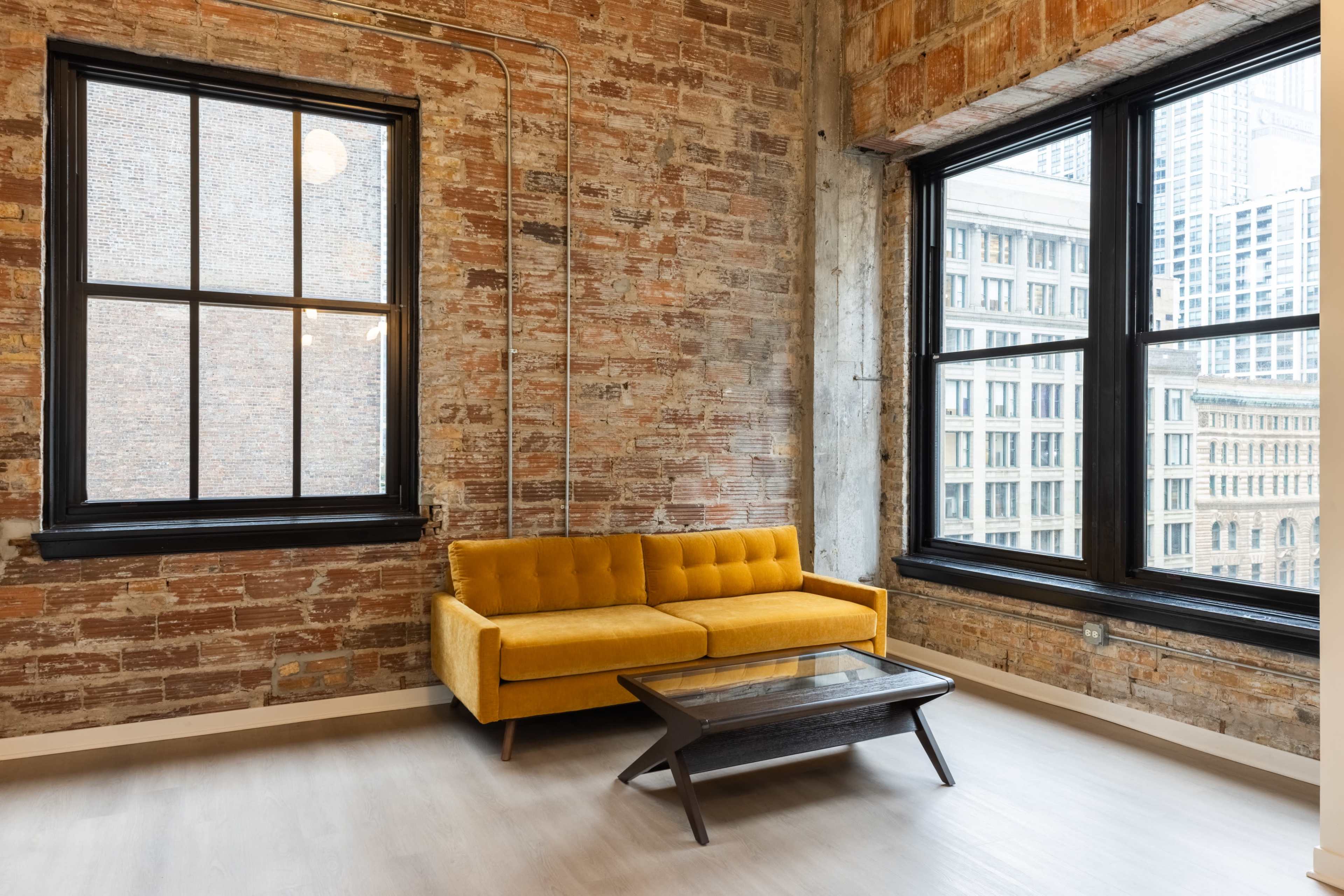 The image shows a modern living room with a yellow sofa and a wooden coffee table set against a backdrop of exposed brick walls and large windows.