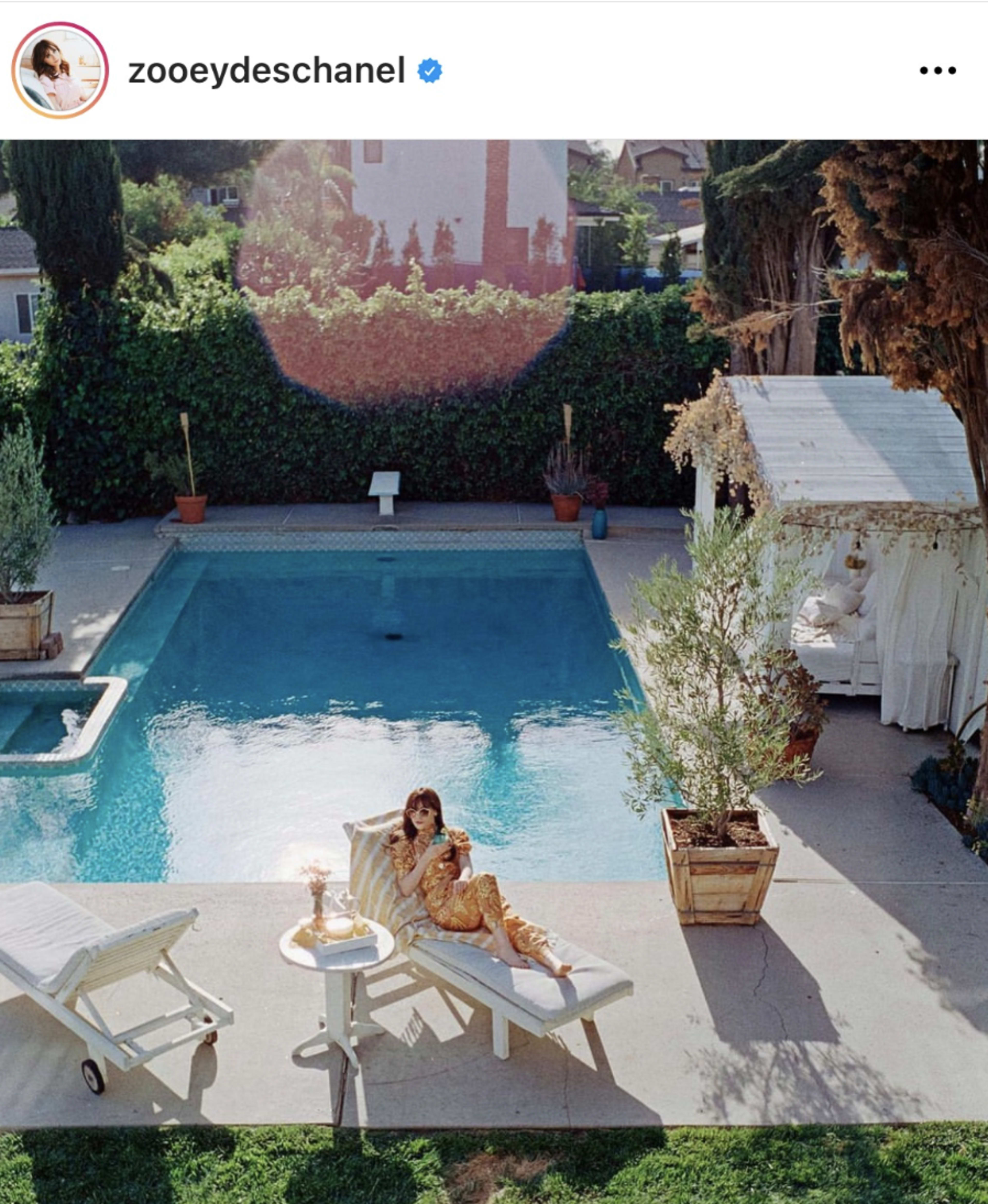 A woman sits on a lounge chair next to a swimming pool surrounded by greenery.