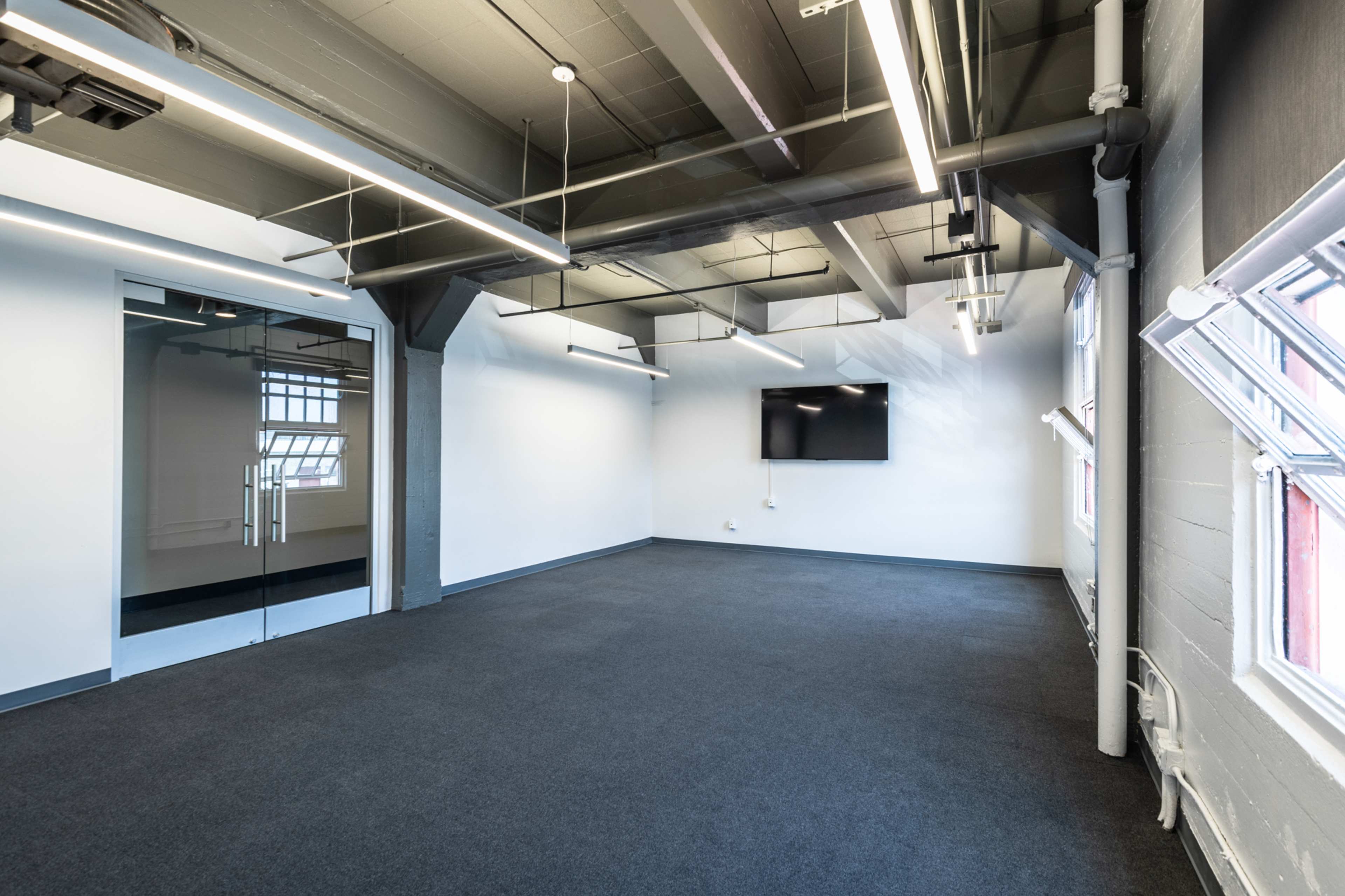 A modern, empty office space with gray carpeting, exposed beams, and a wall-mounted television.