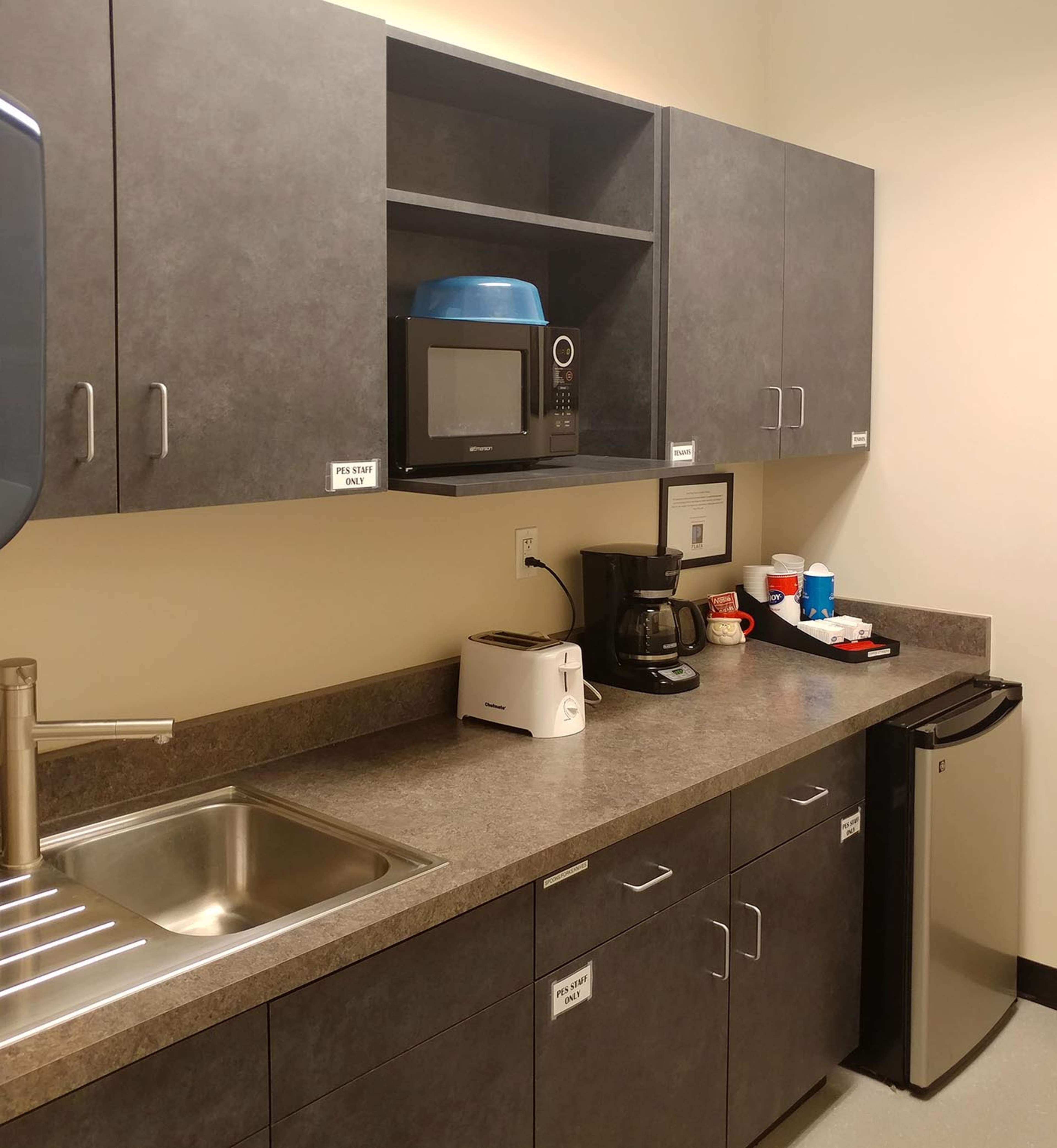 A kitchenette featuring a sink, a microwave, a coffee maker, and a small refrigerator, all against a backdrop of gray cabinets and a countertop.