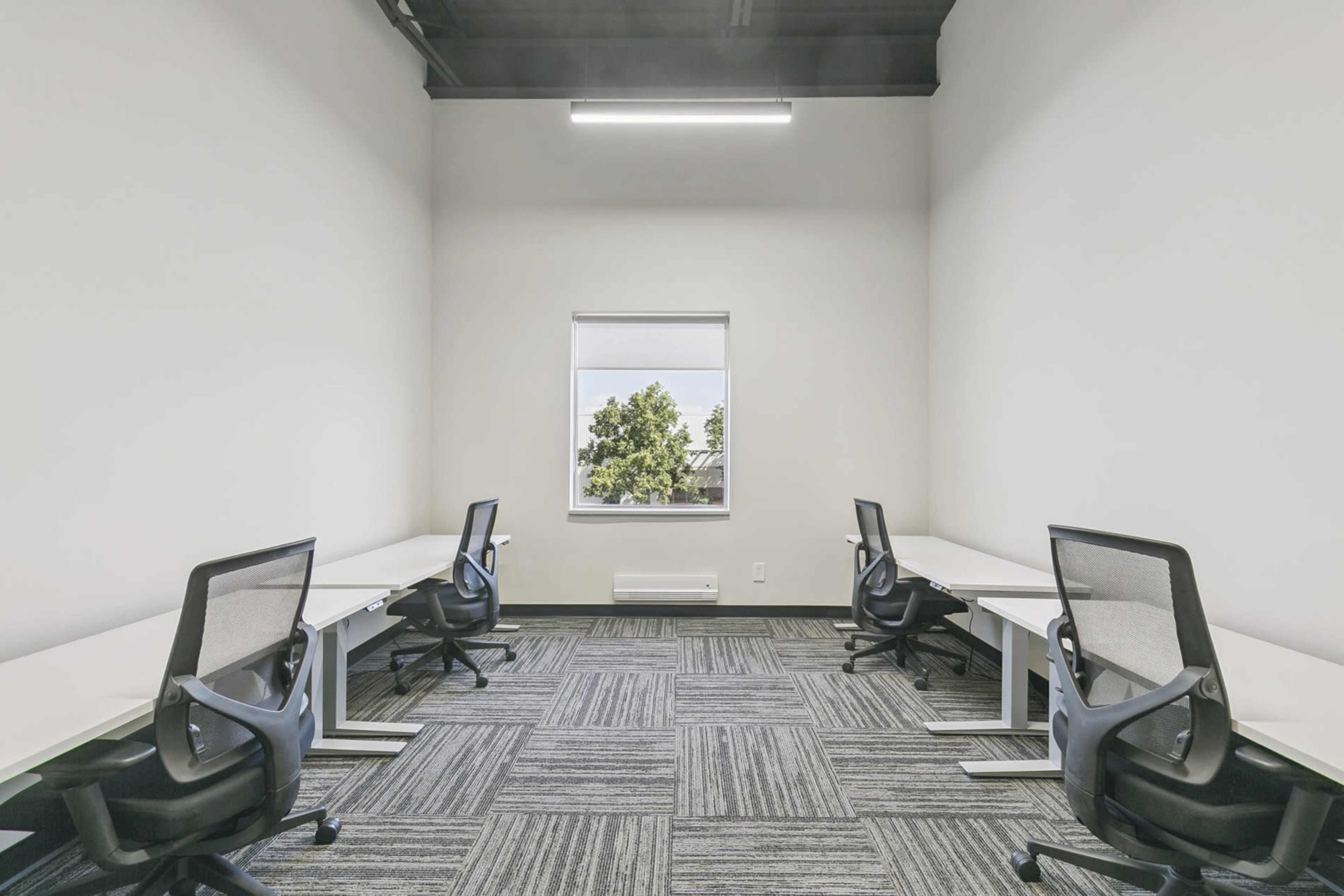 The image shows a vacant office space with four desks and chairs arranged around a window, illuminated by overhead lighting.