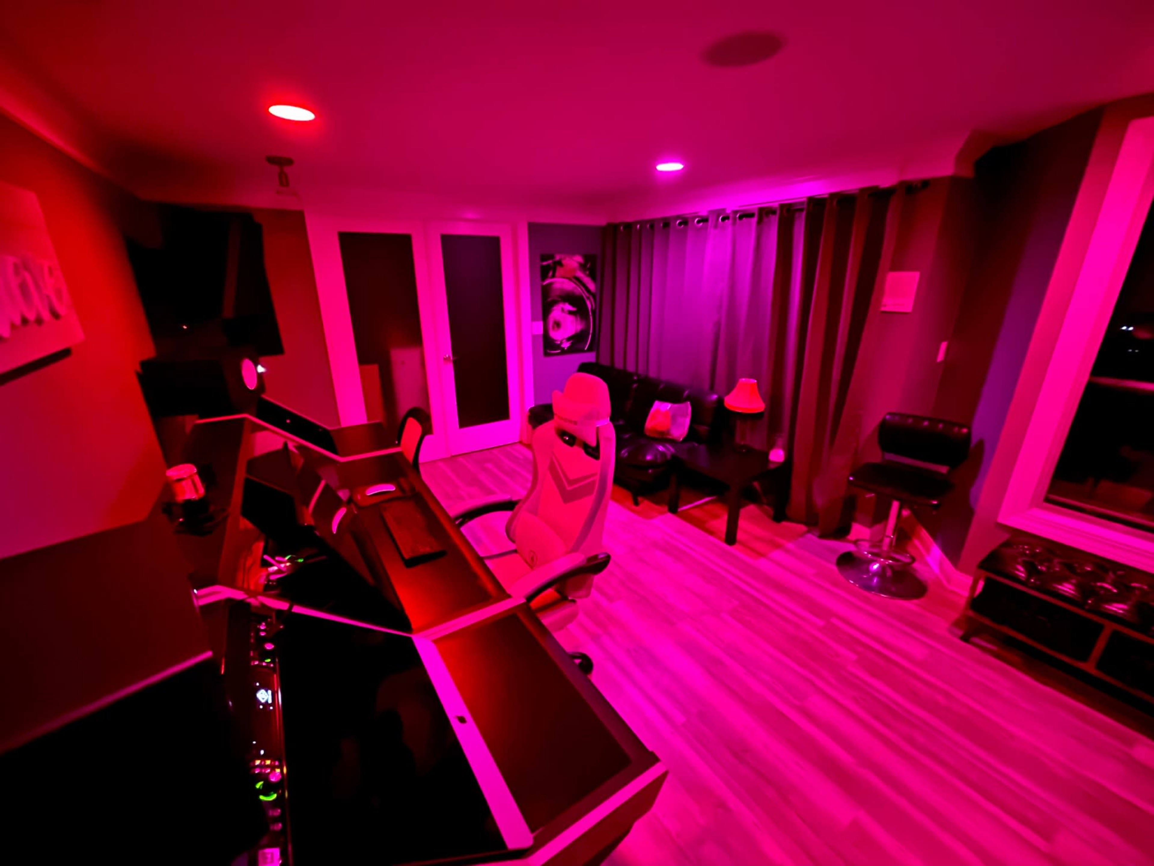 A modern office space features dual computer desks, a gaming chair, and ambient lighting in shades of pink.