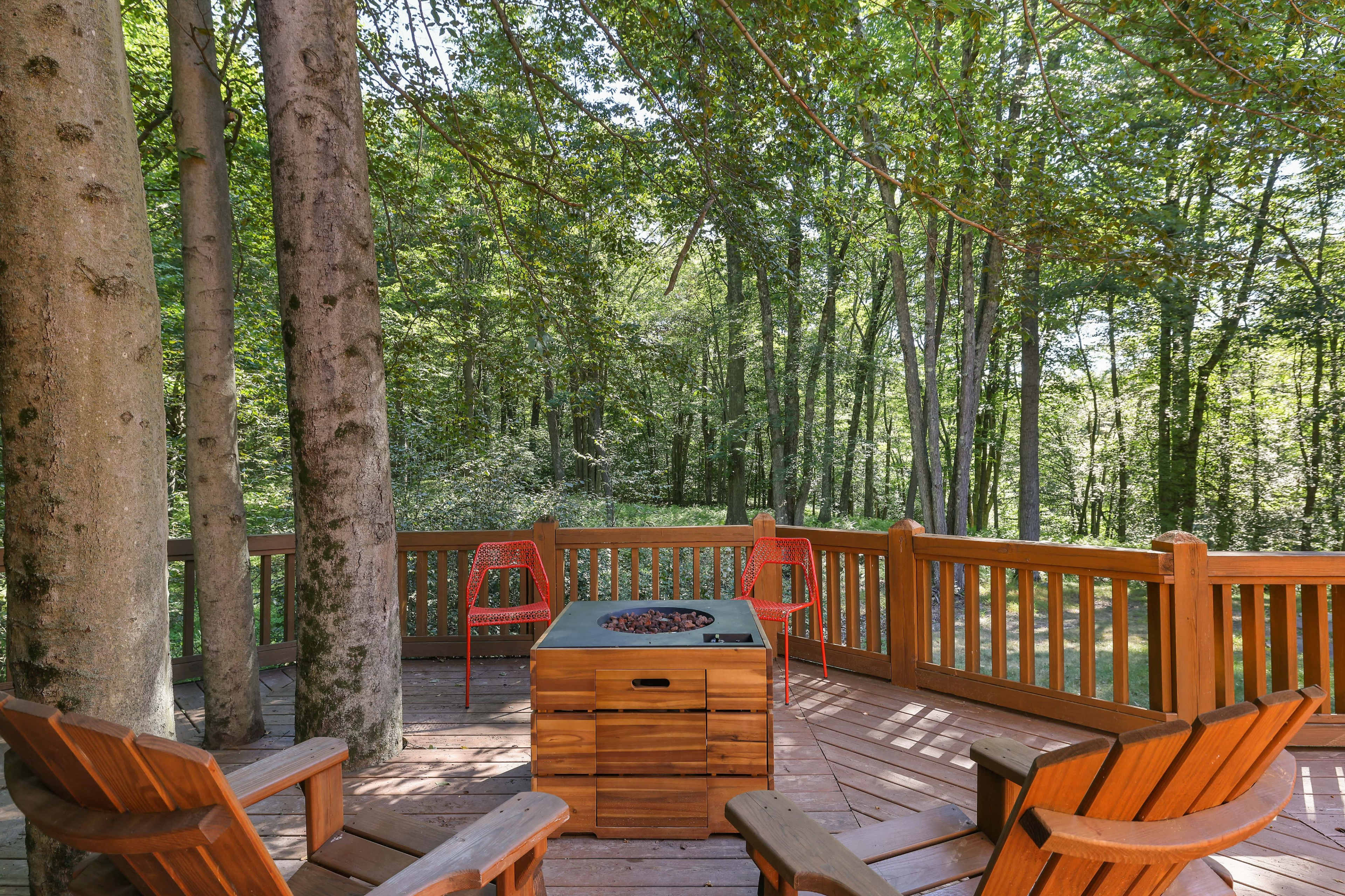 A wooden deck nestled among trees features a fire pit surrounded by four Adirondack chairs and two red metal chairs.