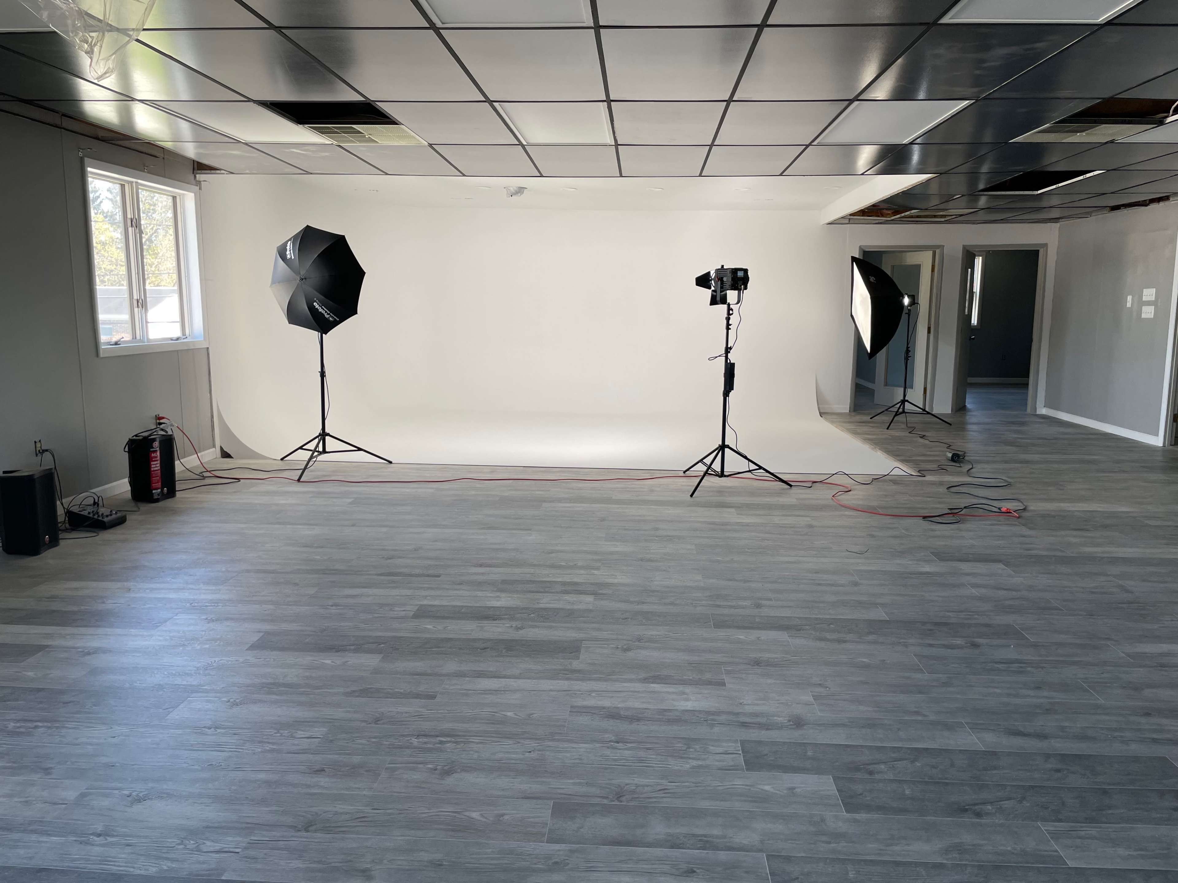 A spacious room with a seamless white backdrop, two studio lights on stands, and gray laminate flooring.