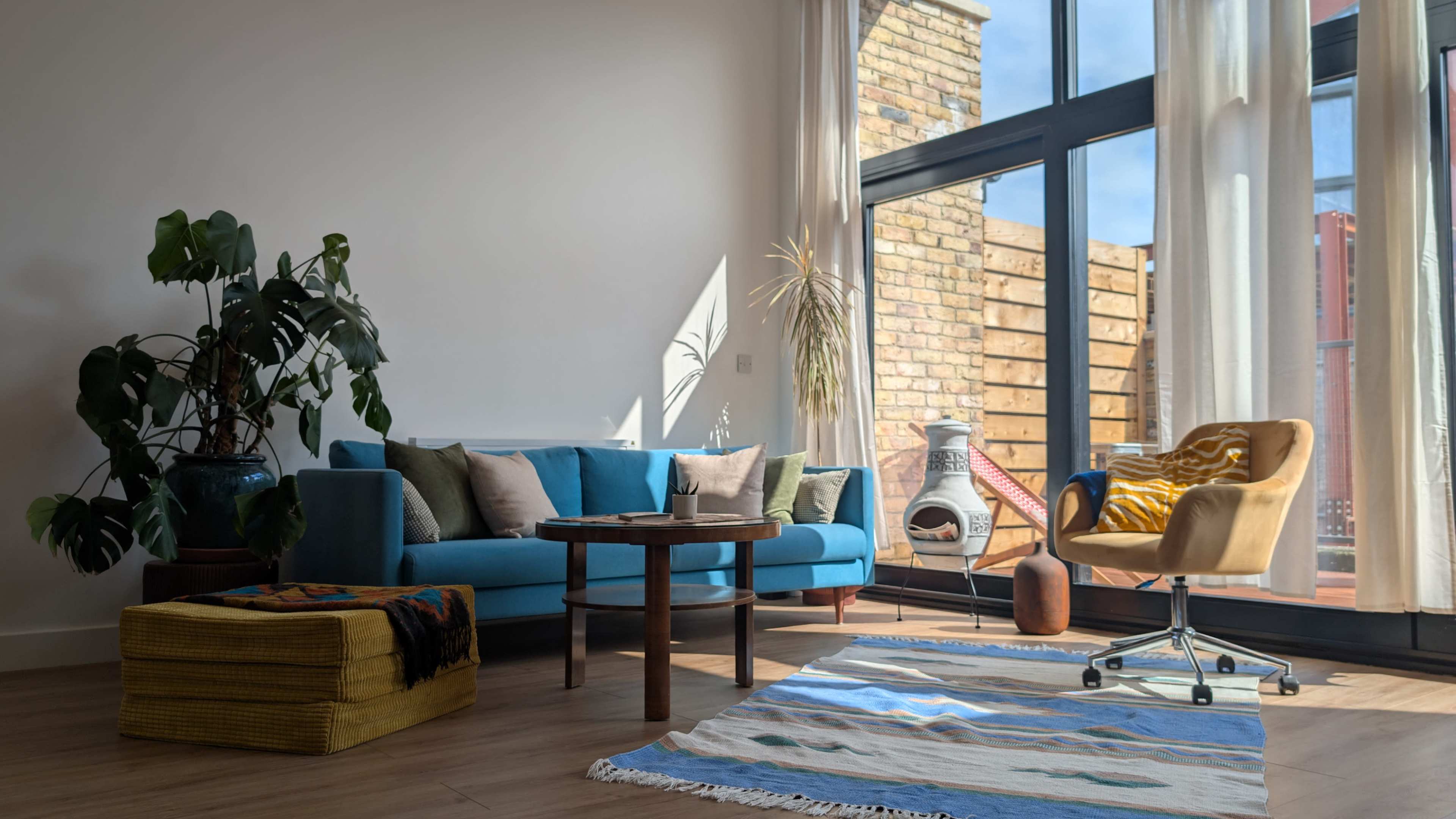 A bright living room features a blue sofa, a round wooden table, a chair, and a patterned rug, with large windows allowing sunlight to enter.