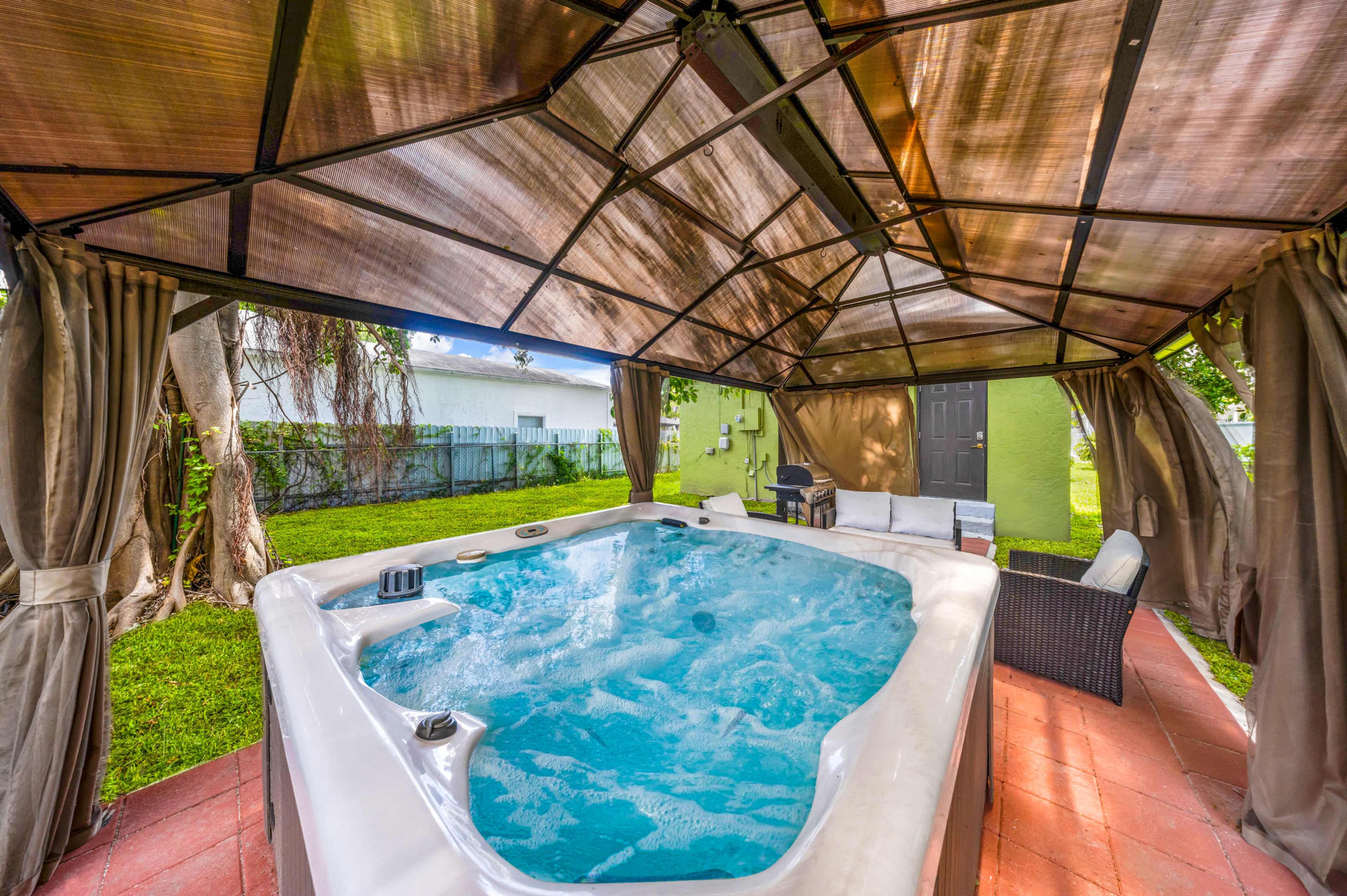 The image shows a hot tub situated under a covered gazebo surrounded by a lawn.