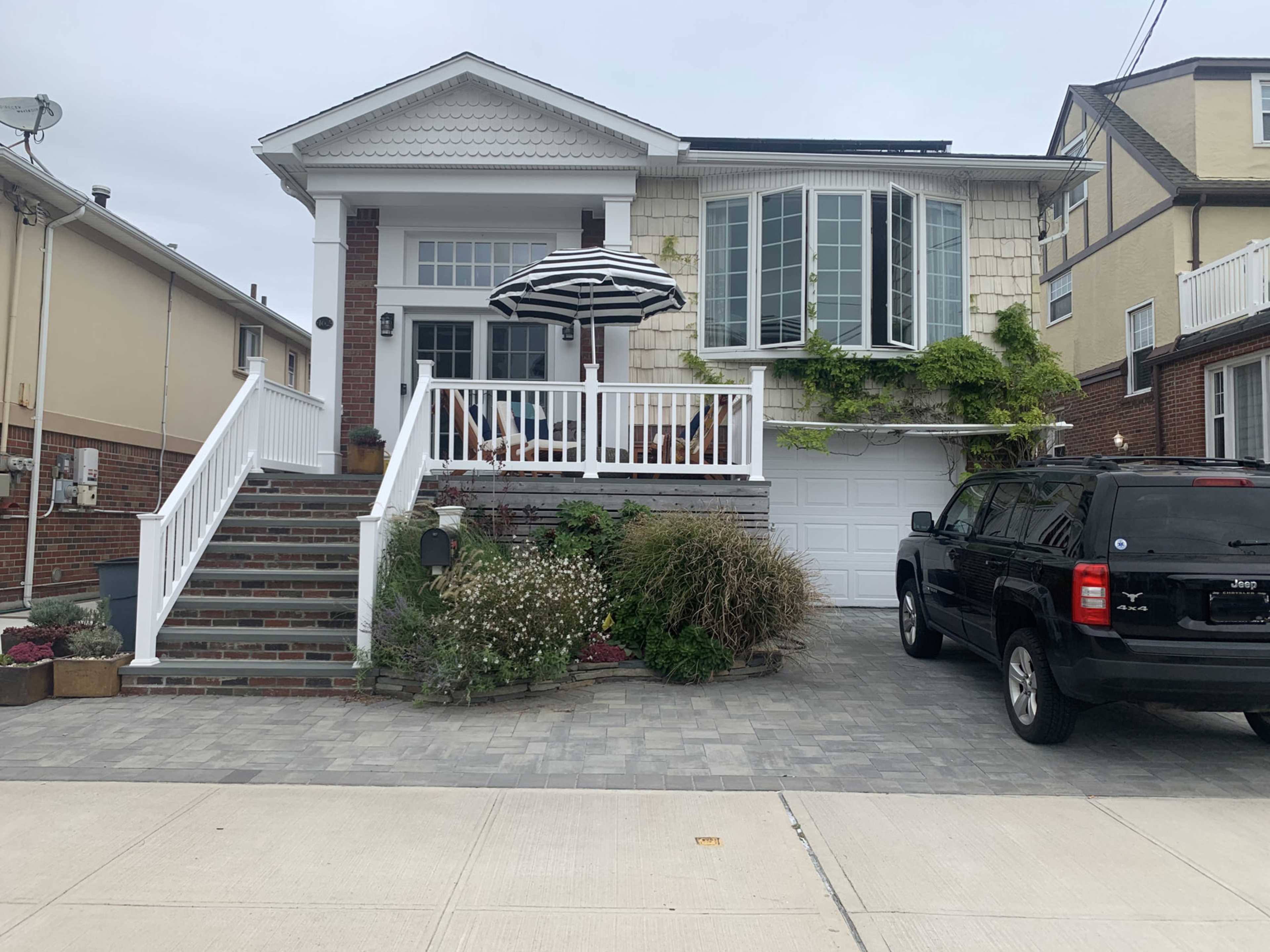 The image shows a two-story house with a front porch, an umbrella, and a landscaped yard, along with a driveway containing a black SUV.