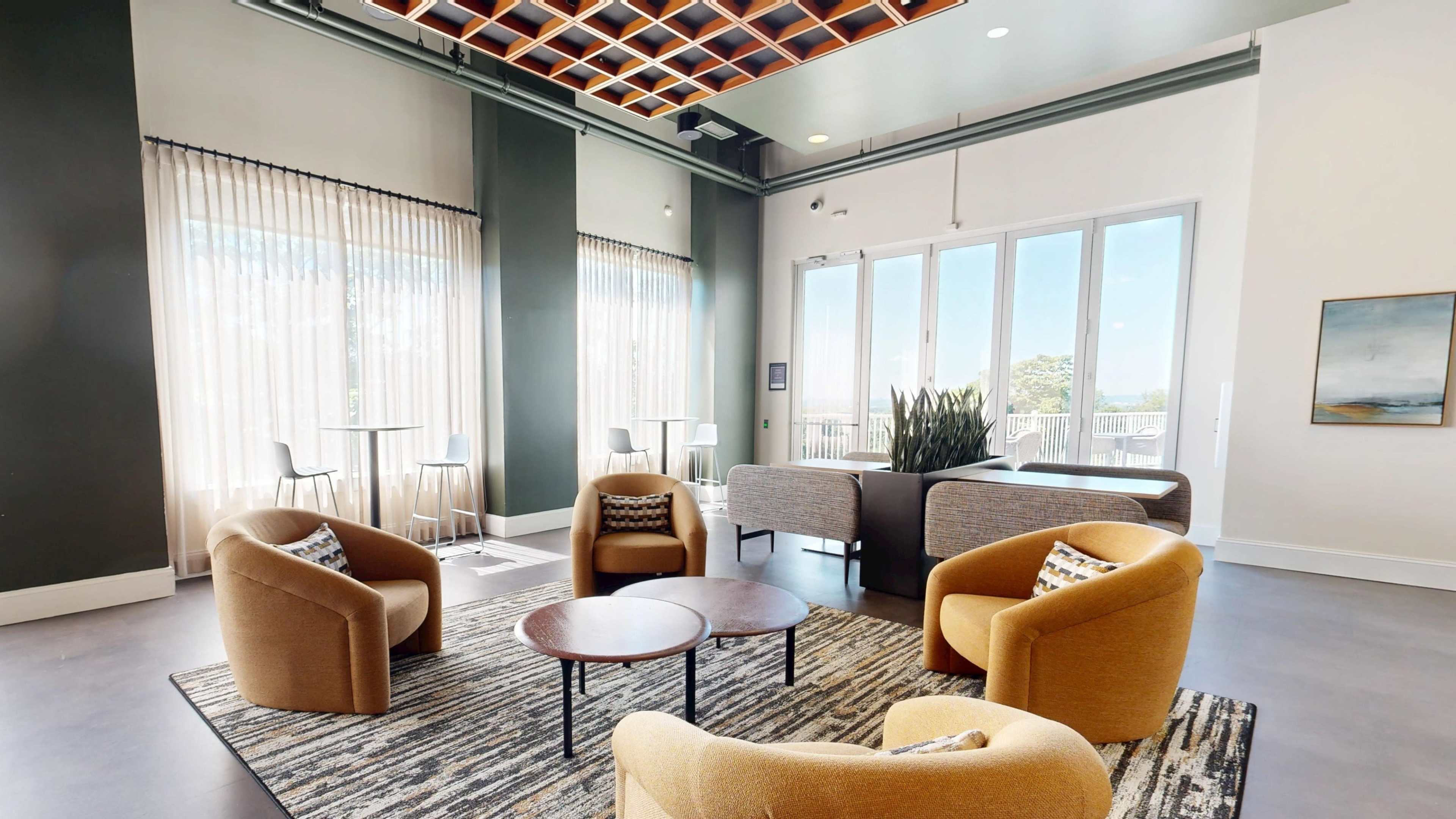 A modern lounge area features four upholstered chairs arranged around a circular table, with large windows allowing natural light to flood the space.