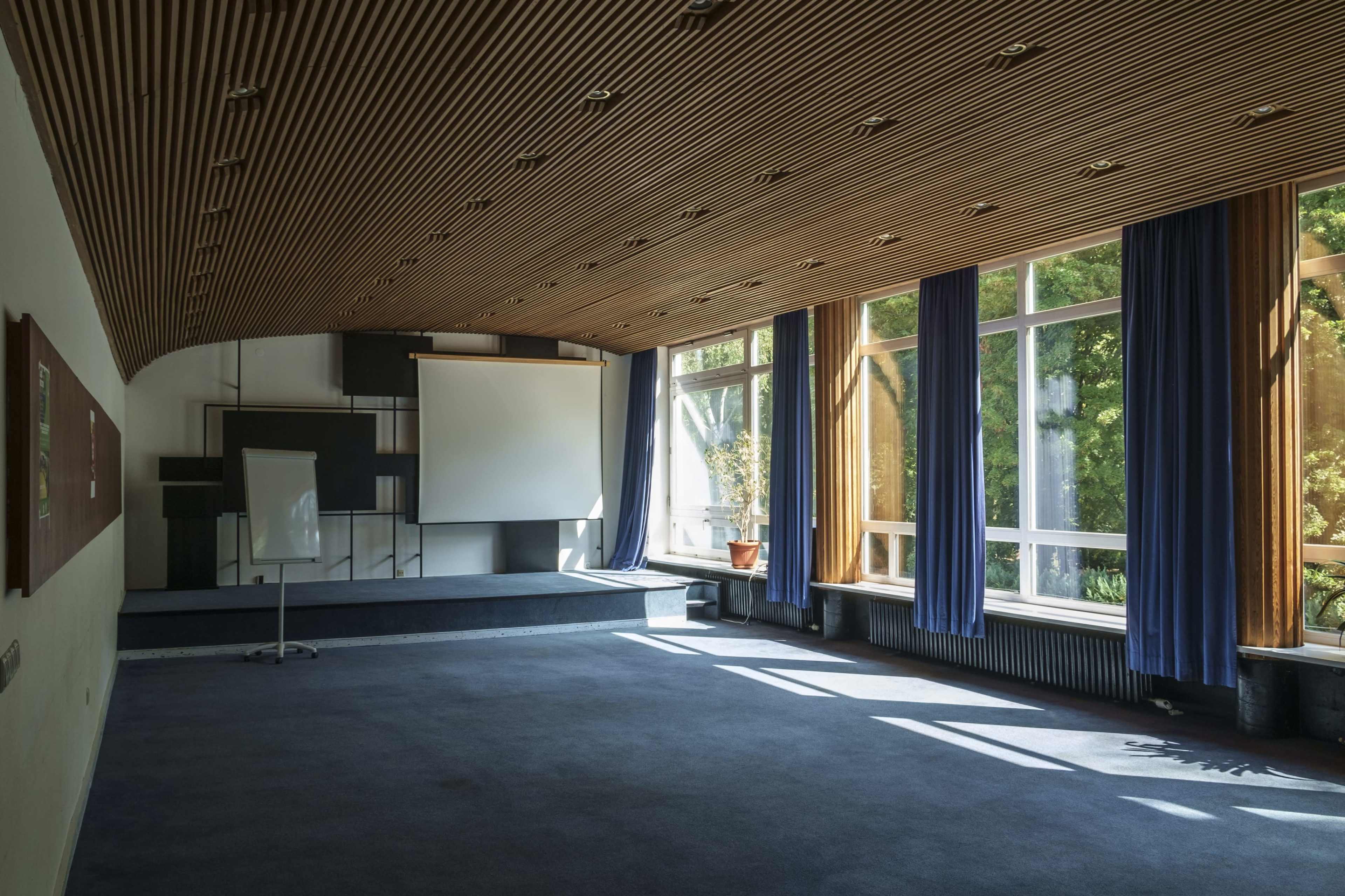 The image shows a spacious conference room with a wooden ceiling, large windows allowing natural light, and a projector screen at the front.