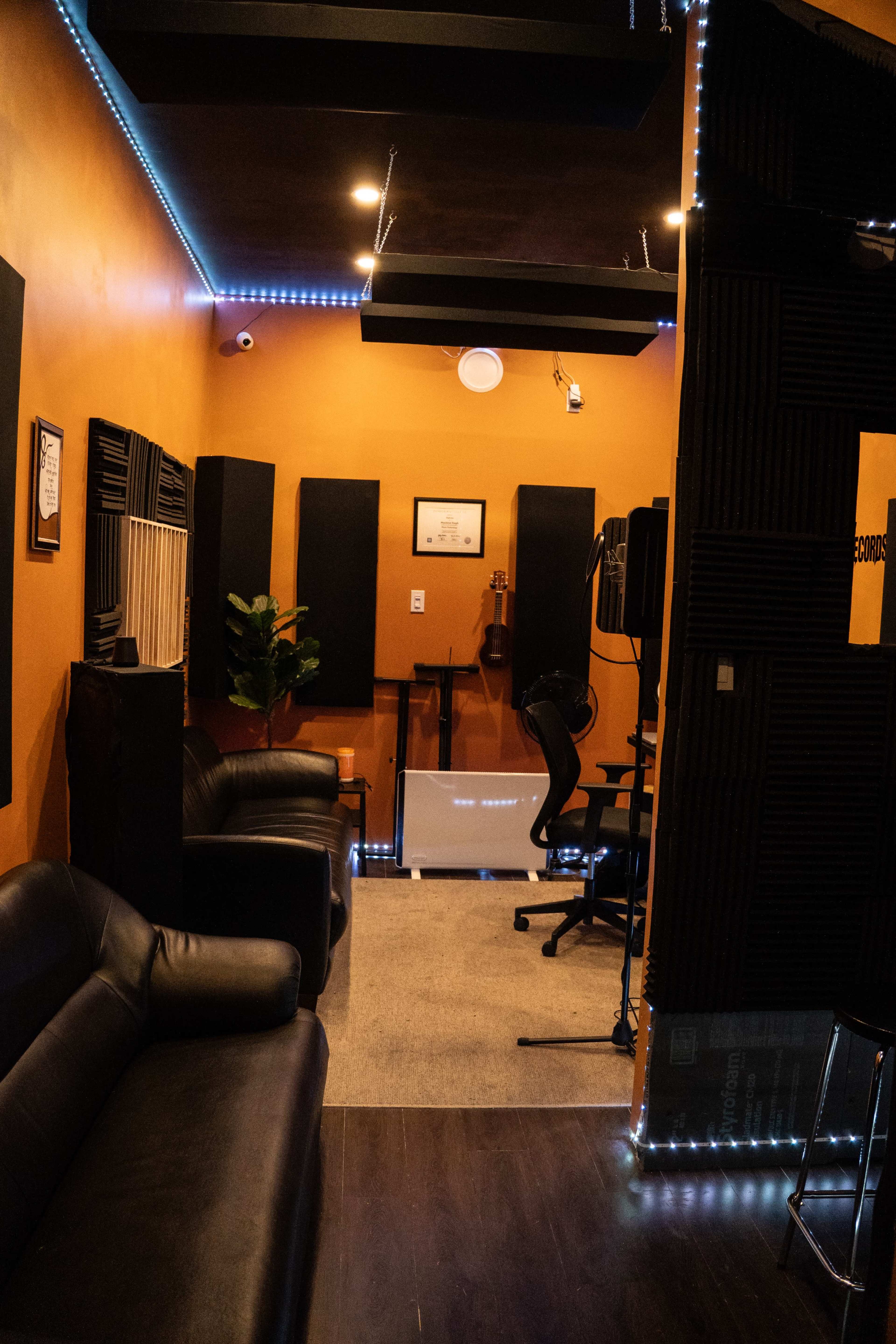 The image shows a recording studio with dark orange walls, black soundproofing panels, seating, and equipment arranged for music production.