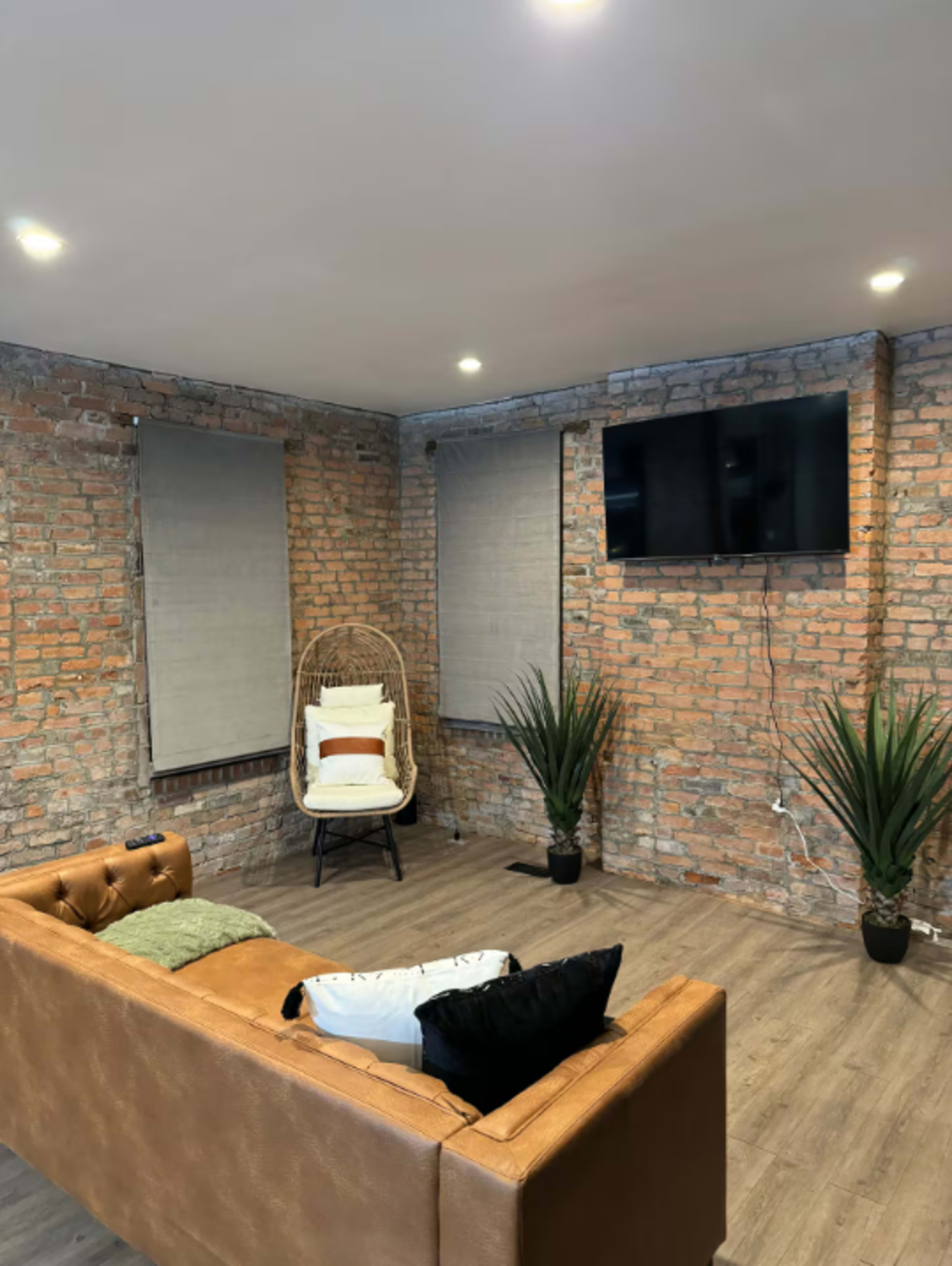 The image shows a cozy room with exposed brick walls, a brown leather sofa, a hanging chair, and a wall-mounted TV.
