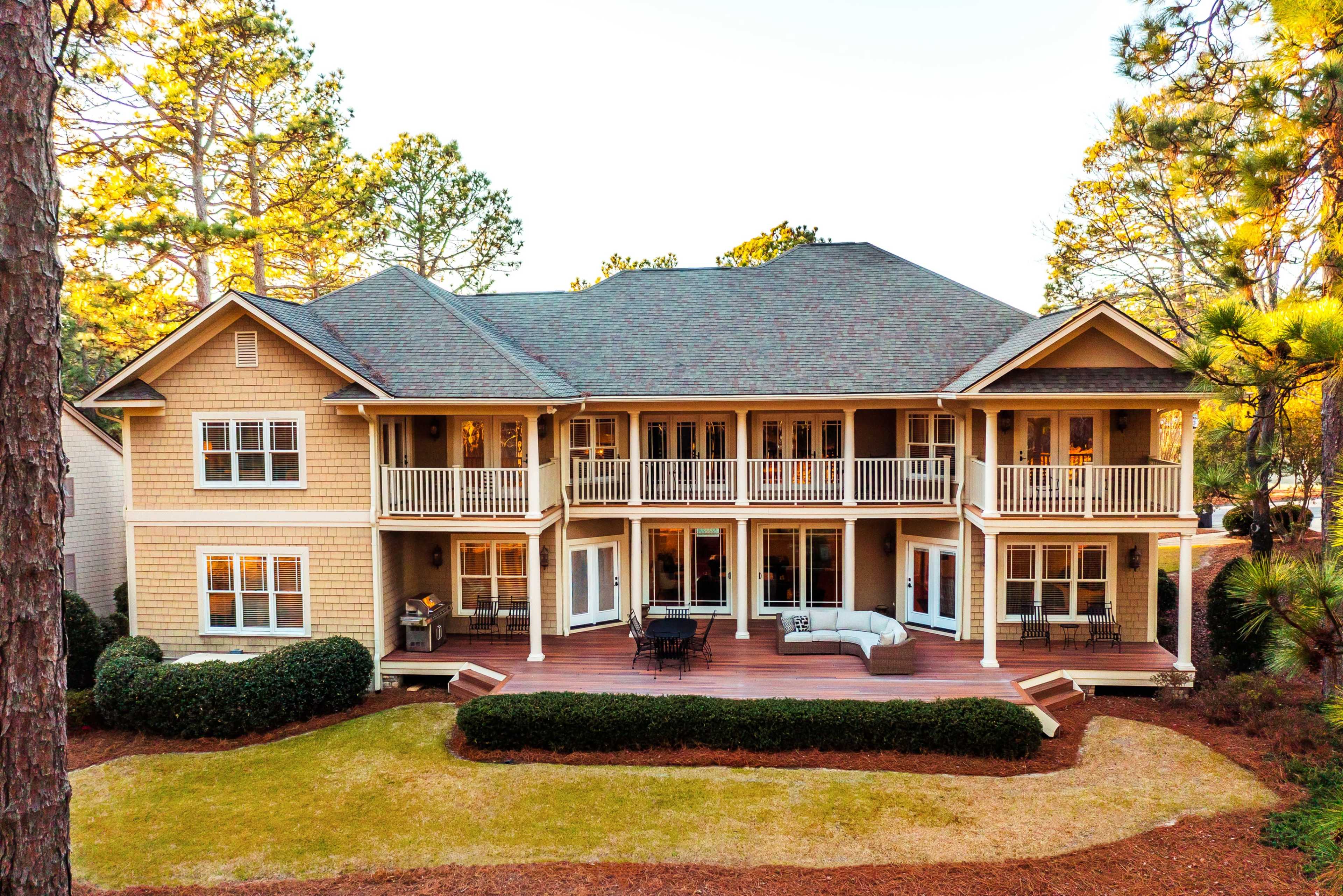 A large two-story house with multiple balconies and a wooden deck overlooks a landscaped yard surrounded by pine trees.
