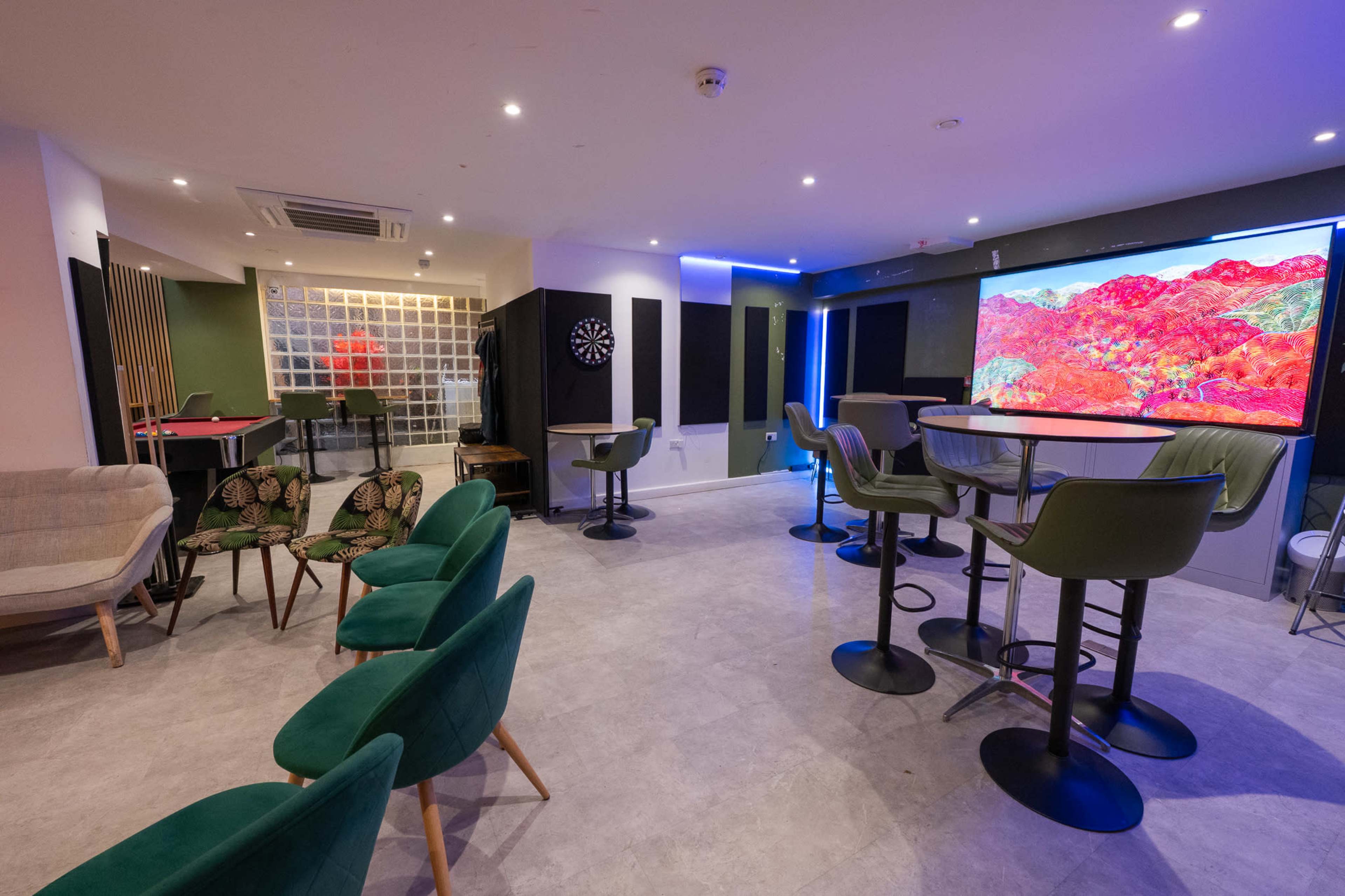 The image shows a modern lounge area with seating, a dartboard, and a large screen displaying colorful artwork.